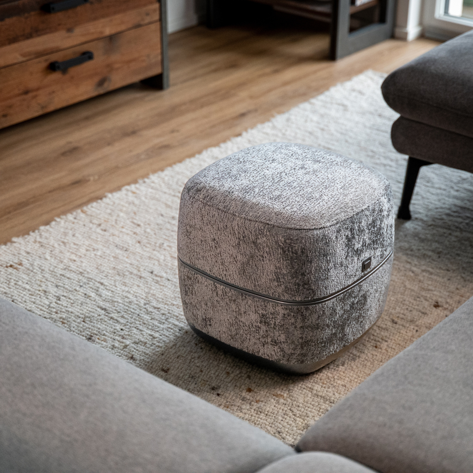 Ein grauer Hocker mit Muster steht auf einem Teppich neben einem Sofa.
