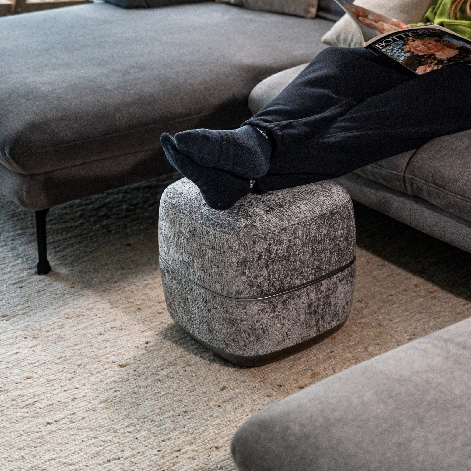 Füße in schwarzen Socken auf grauem Hocker vor grauem Sofa, liest Magazin.