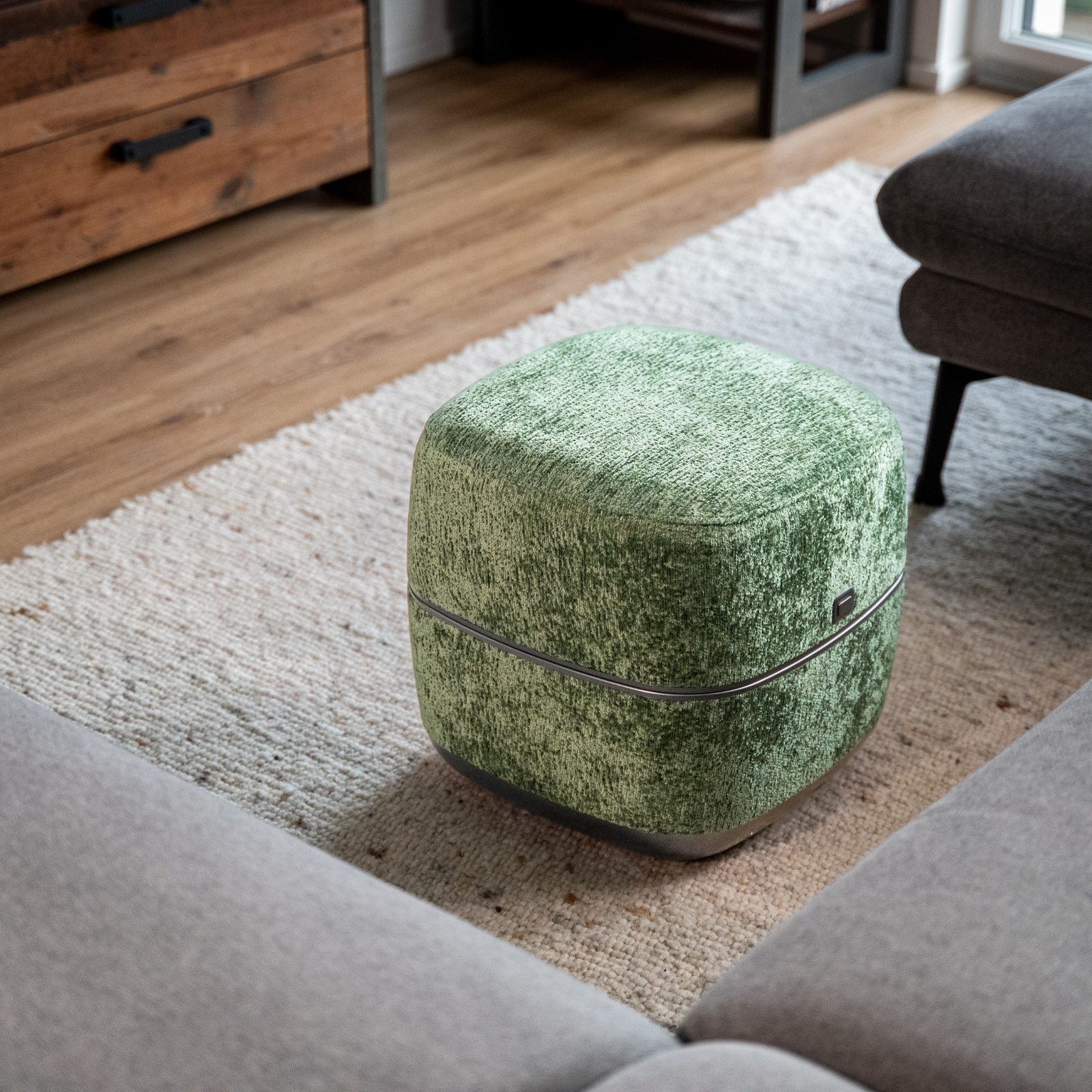 Ein grüner Hocker mit samtiger Textur steht auf einem hellen Teppich neben grauen Sofas.
