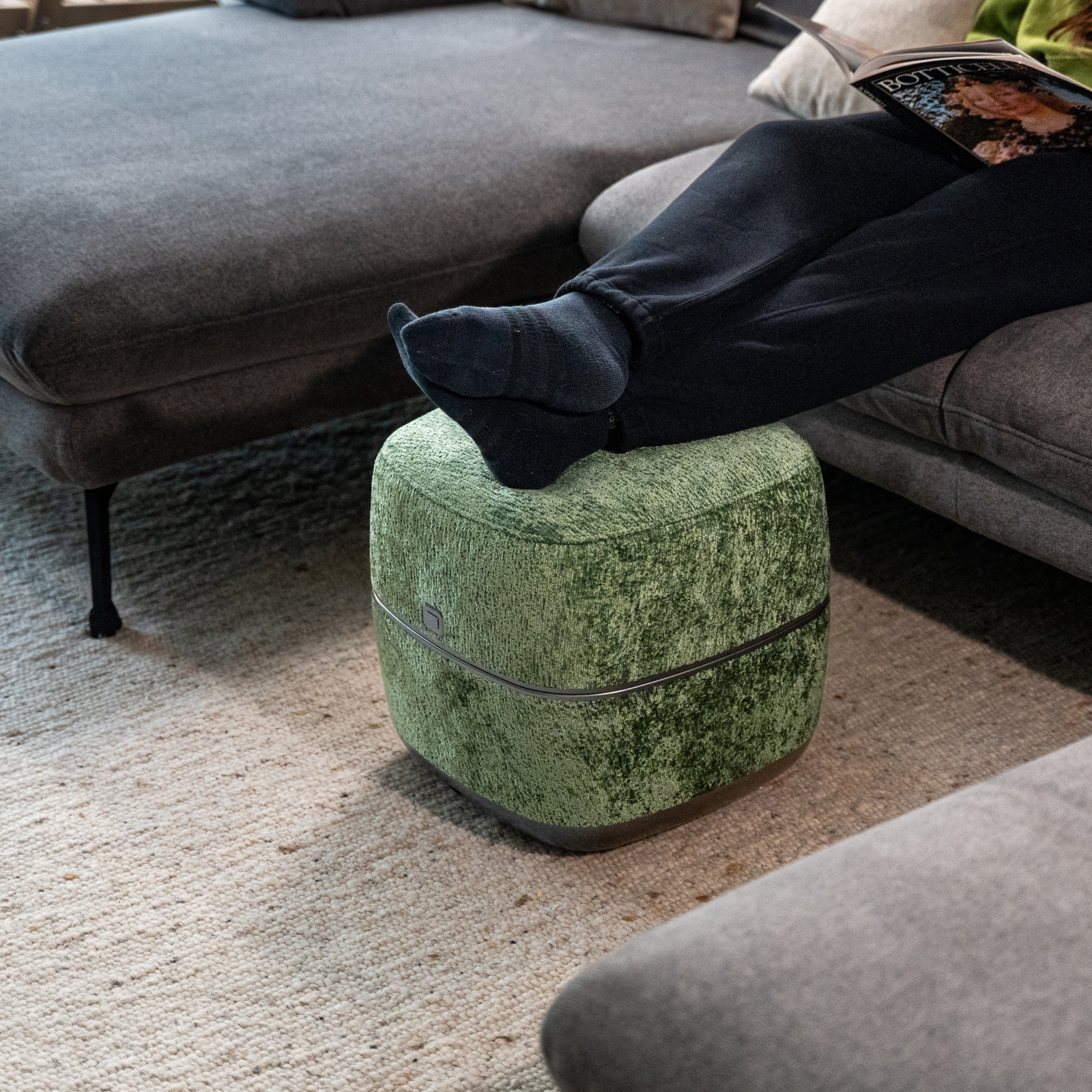 Person mit schwarzen Socken ruht Füße auf einem grünen Hocker neben Sofa.