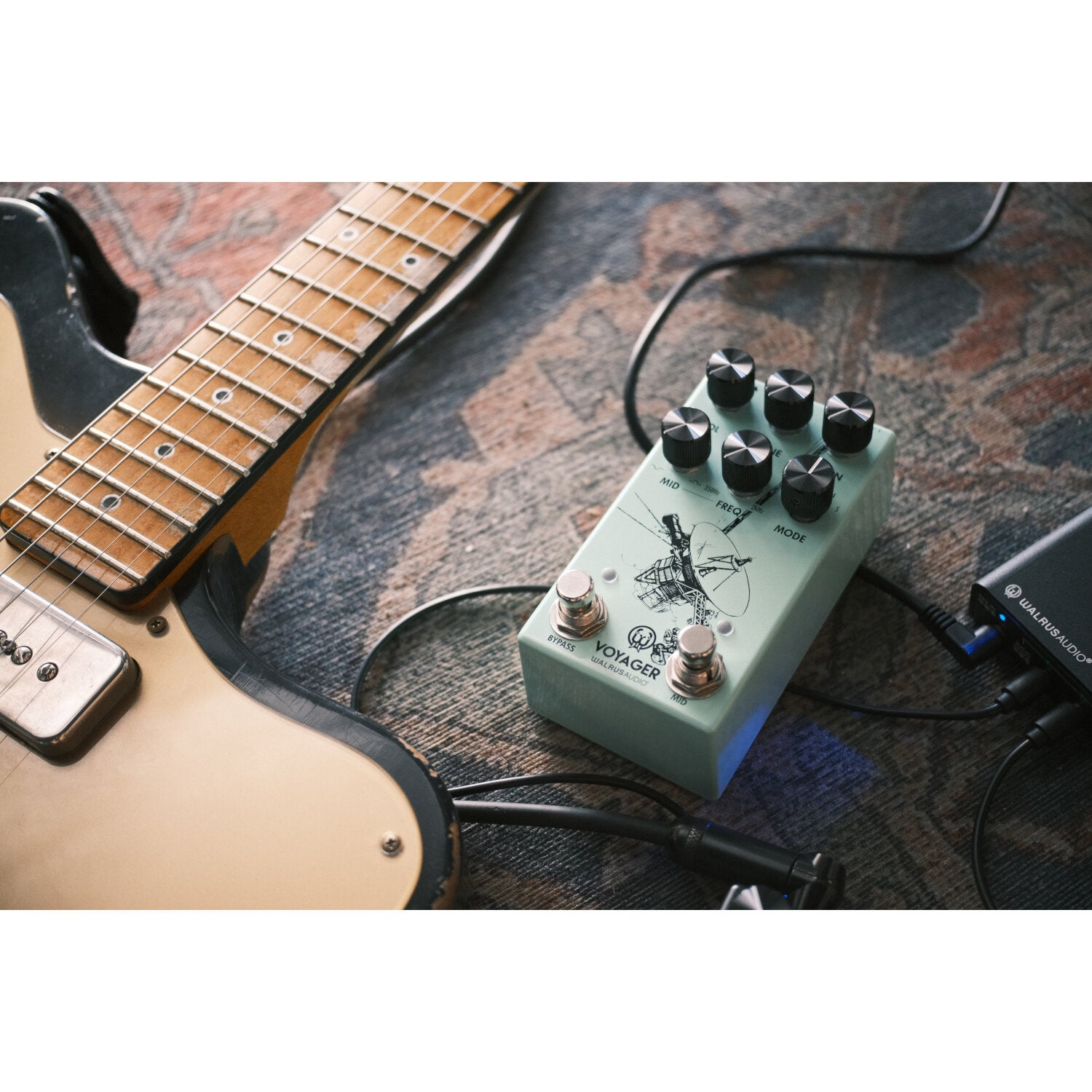A light green guitar pedal sits next to a tan electric guitar on a patterned rug.
