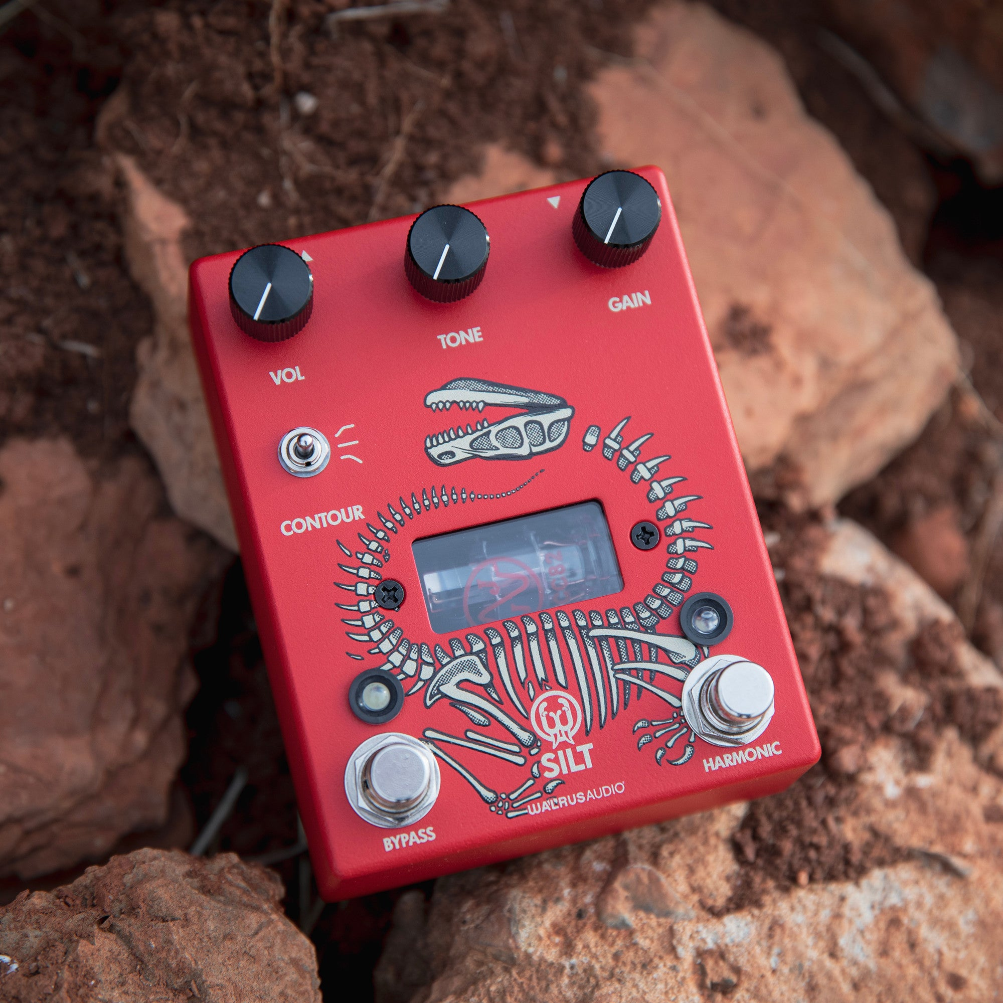 Red guitar effects pedal with dinosaur skeleton design, resting on rocks.