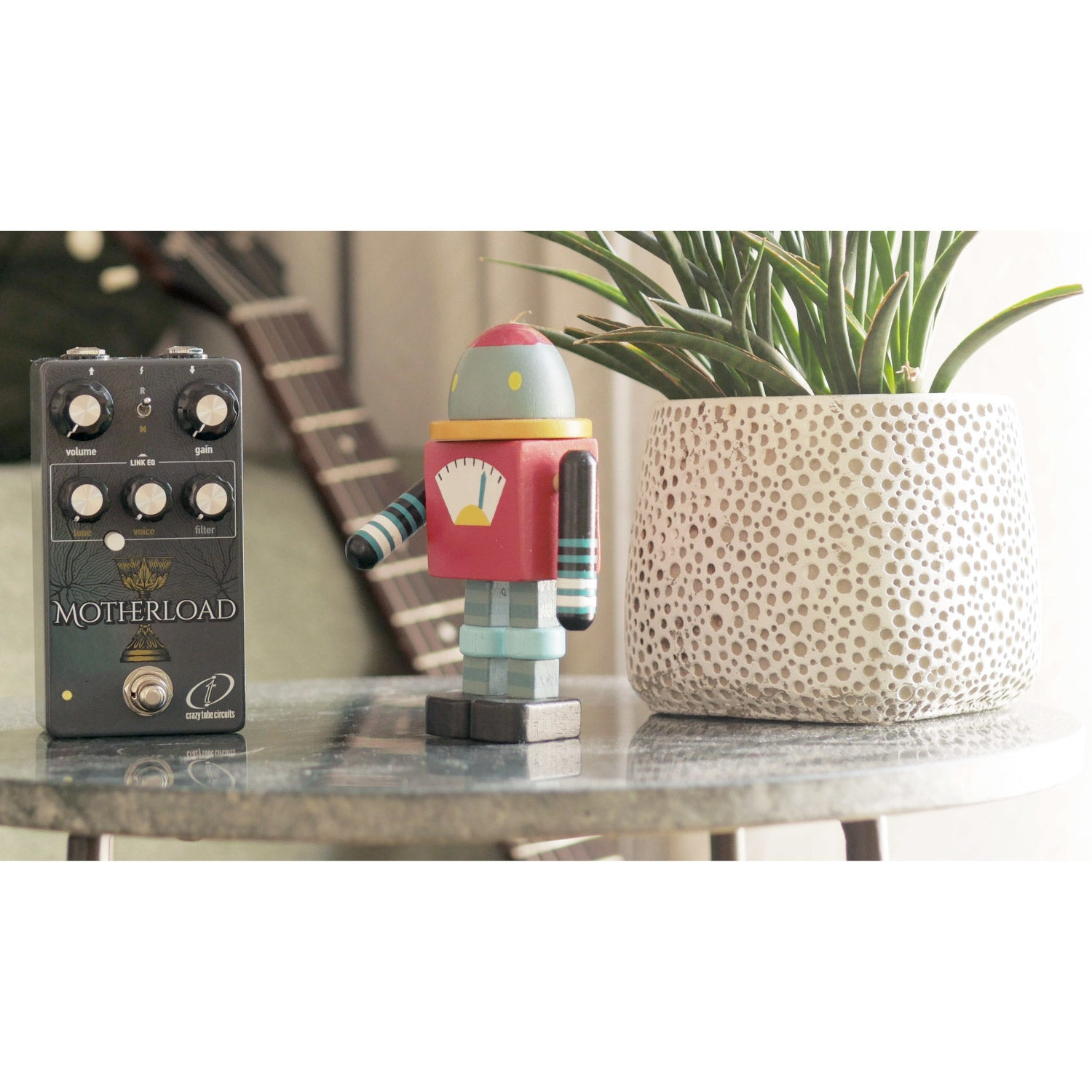 A guitar pedal, wooden robot, and potted plant on a table.