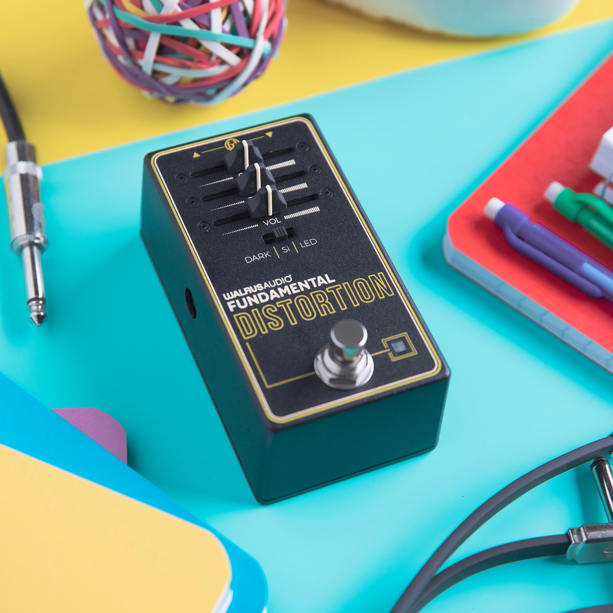 Black distortion pedal with sliders and knobs, on a cyan surface with other items.