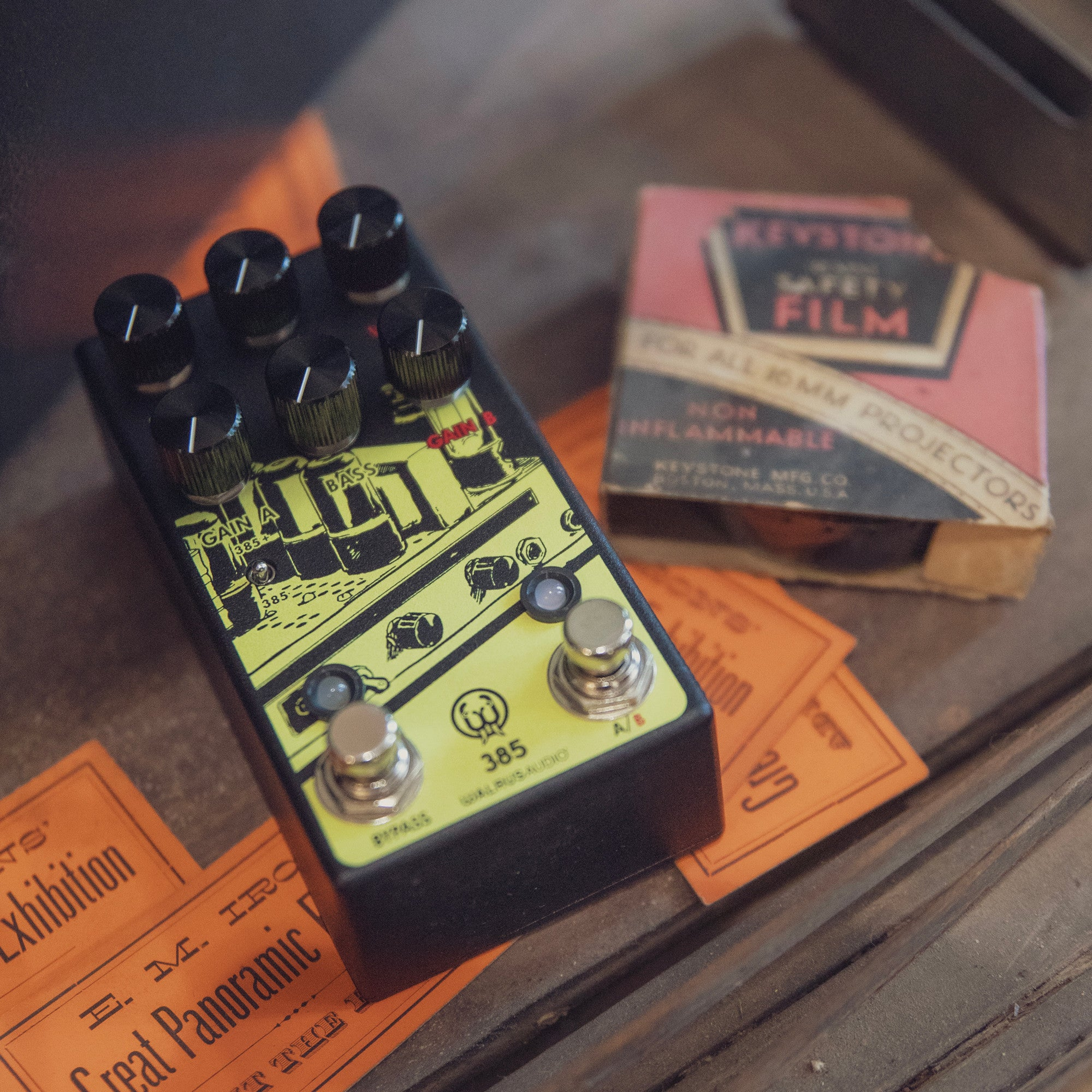 A black guitar pedal with yellow graphics sits on orange tickets and a film box.