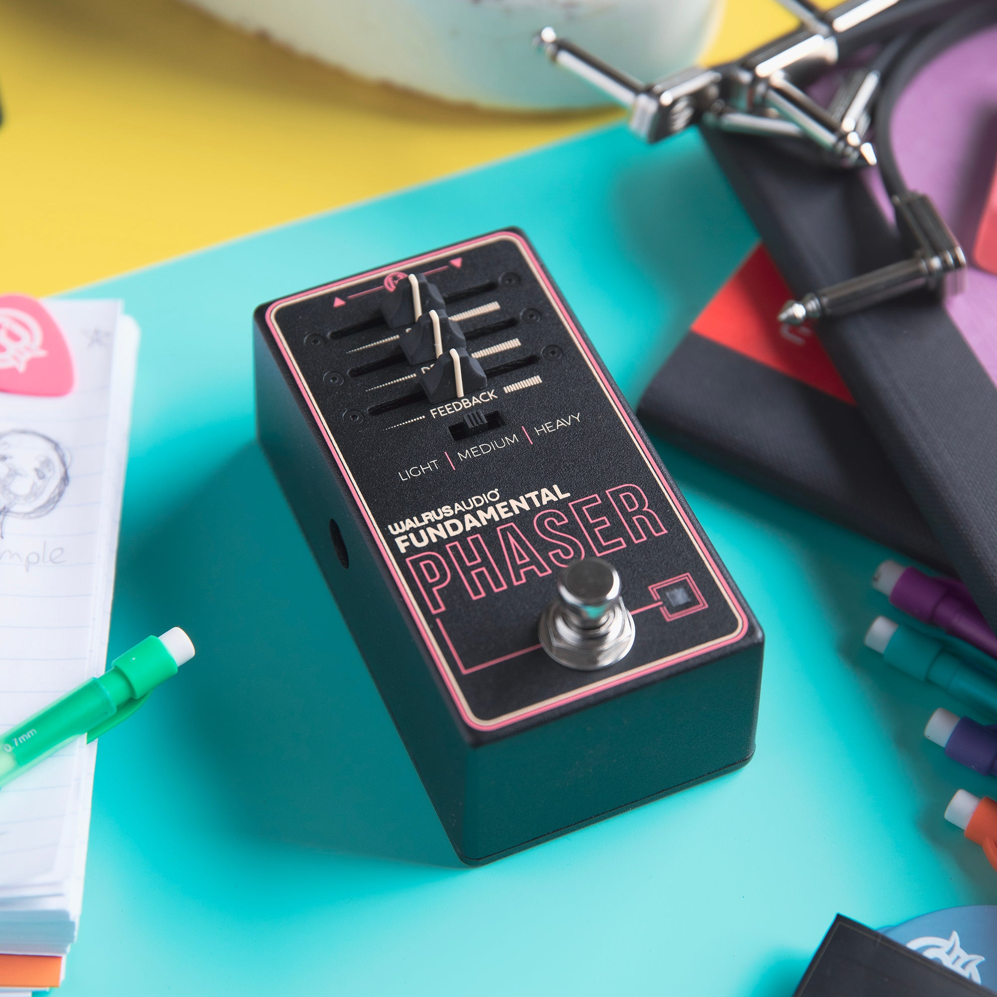 A black phaser pedal with a pink border rests on a turquoise desk.
