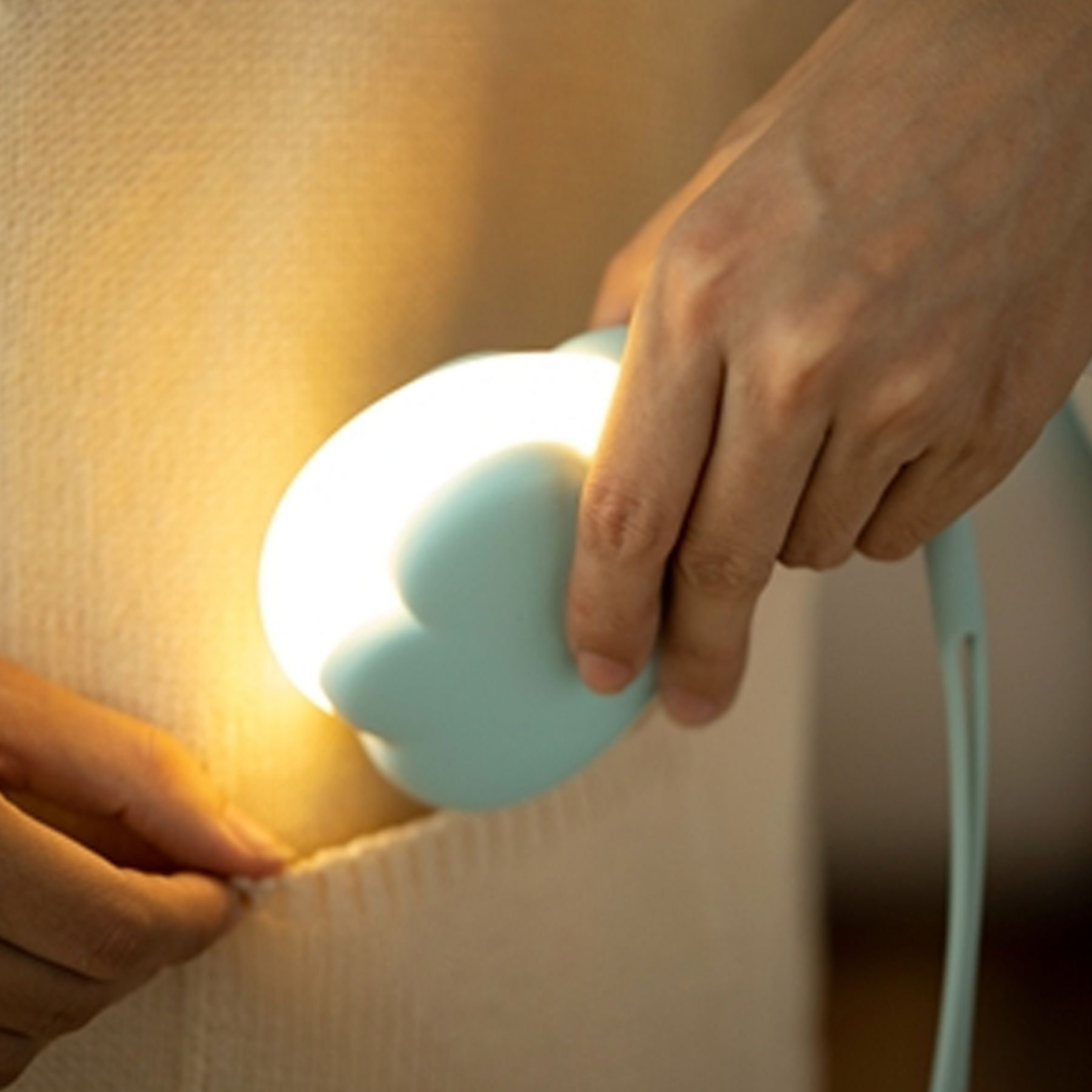 Eine Hand hält eine hellblaue Lampe mit leuchtender Glühbirne über einem beigen Stoff.