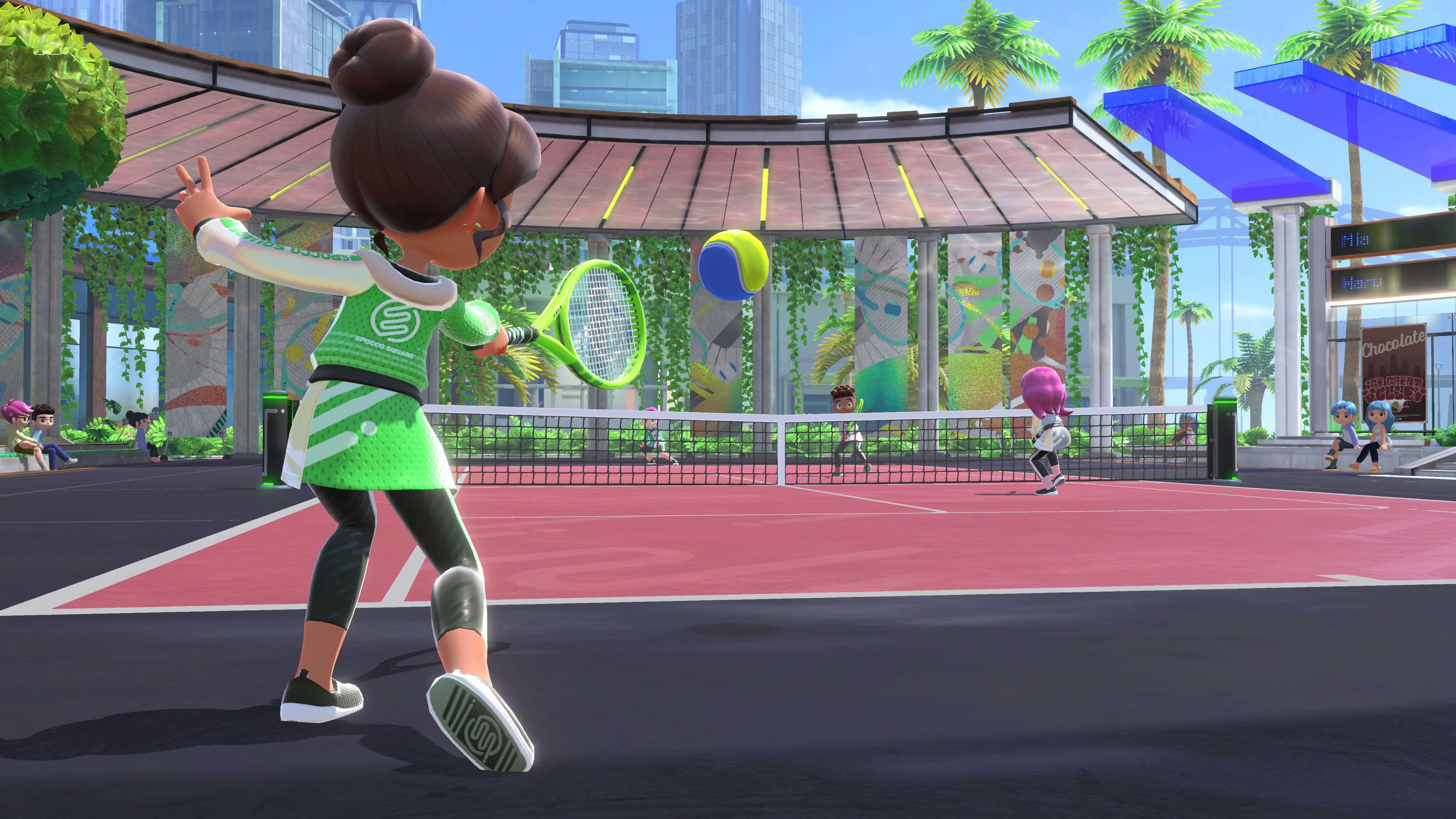 Spieler schlägt Tennisball auf einem roten Platz unter einem Glasdach mit Palmen und Gebäuden im Hintergrund.