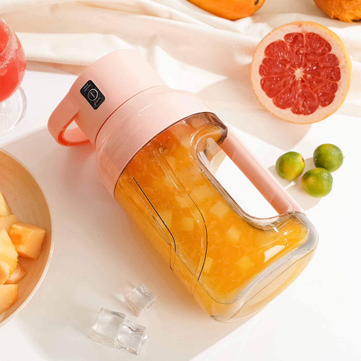 Una licuadora rosa con jugo de naranja, junto a frutas y cubitos de hielo.
