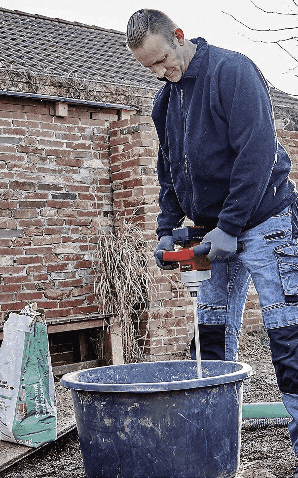 Mann mischt Material in Tonne mit Rührwerk. Ziegelwand und Sack daneben.