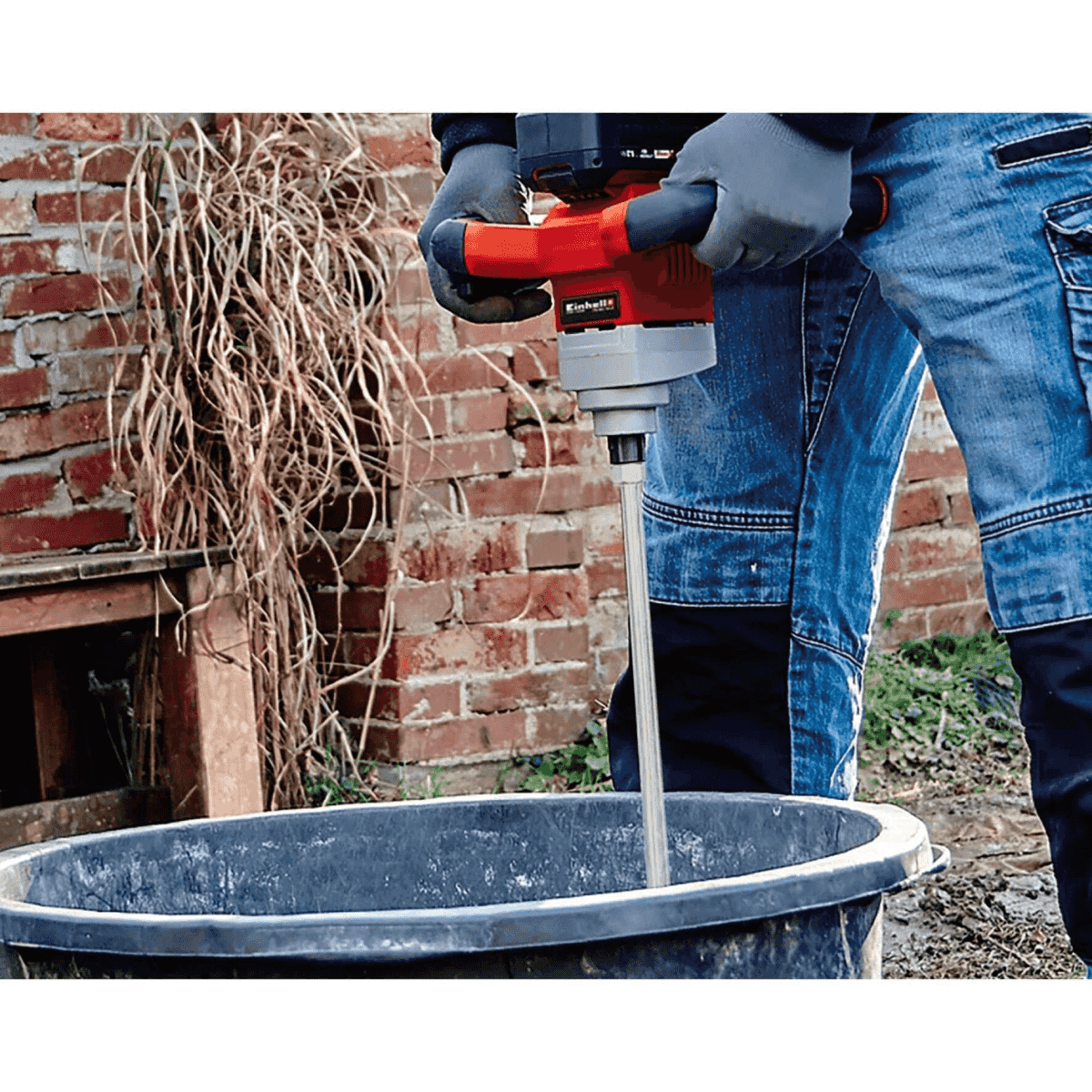 Person mit Rührwerk mischt Mörtel in Eimer vor Backsteinmauer.