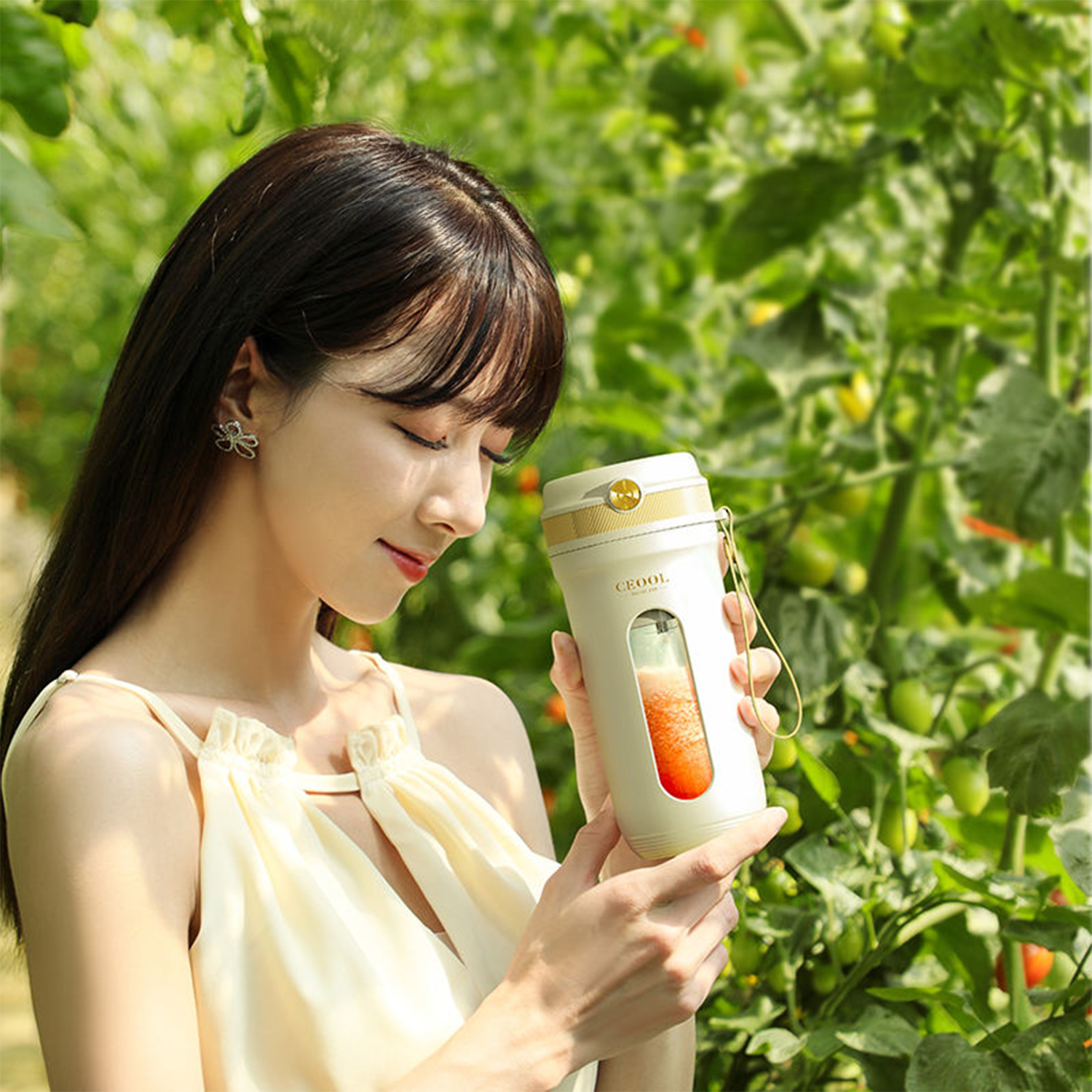 Una mujer con un vestido claro sostiene una licuadora portátil con jugo de naranja en un invernadero.