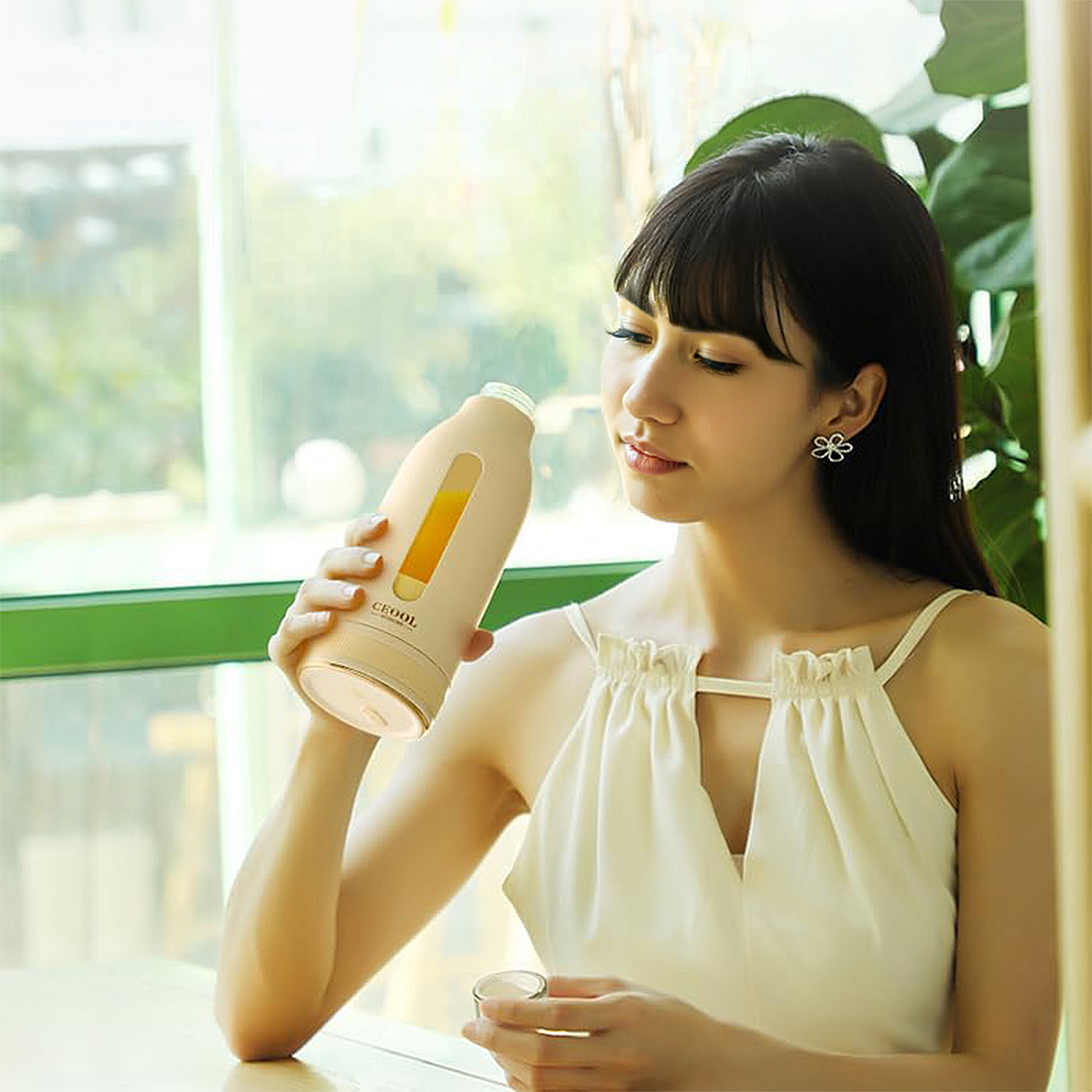 Una mujer sostiene una botella beige con jugo de naranja cerca de una ventana.