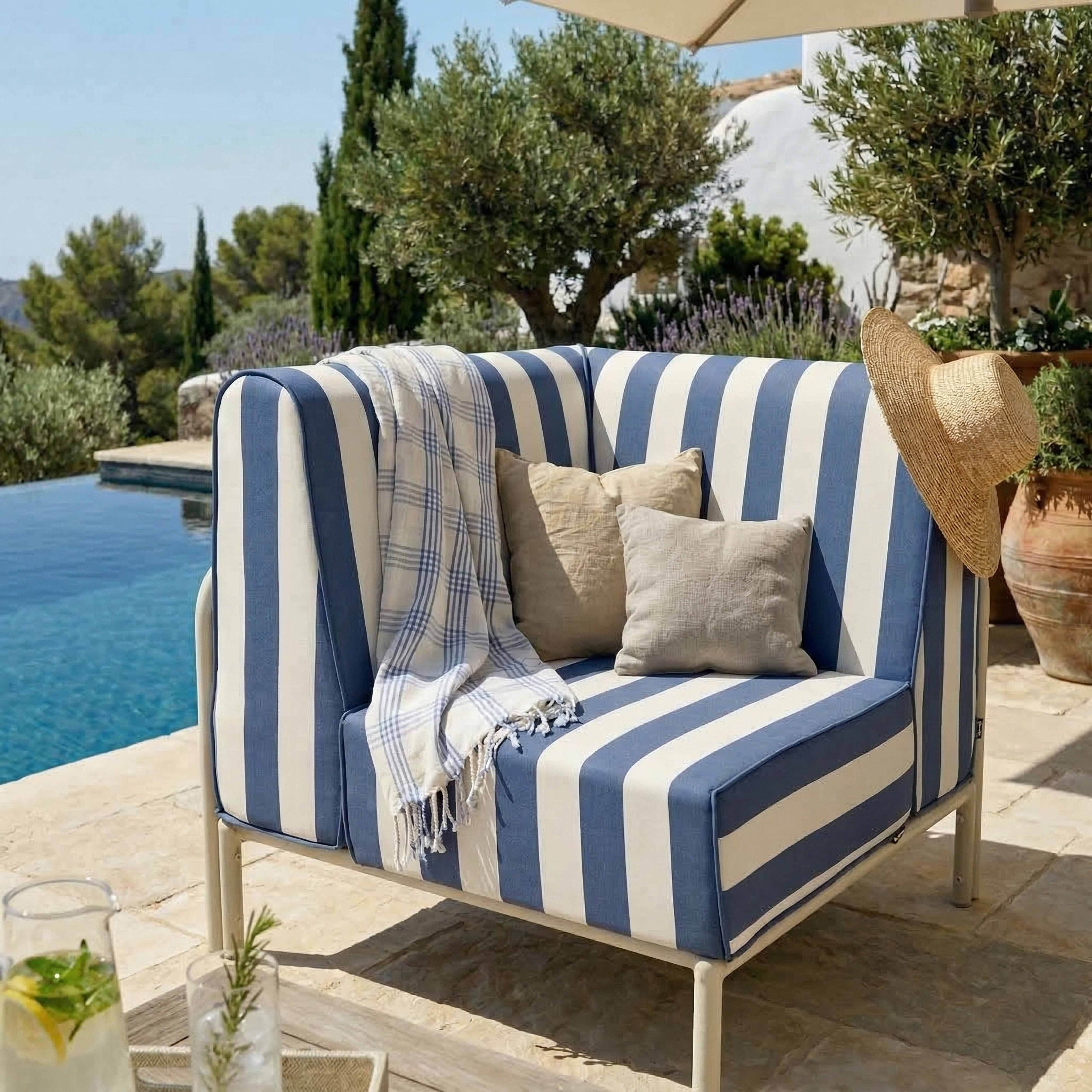 Ein blau-weiß gestreiftes Outdoor-Sofa mit Kissen, Decke und Strohhut neben einem Pool.