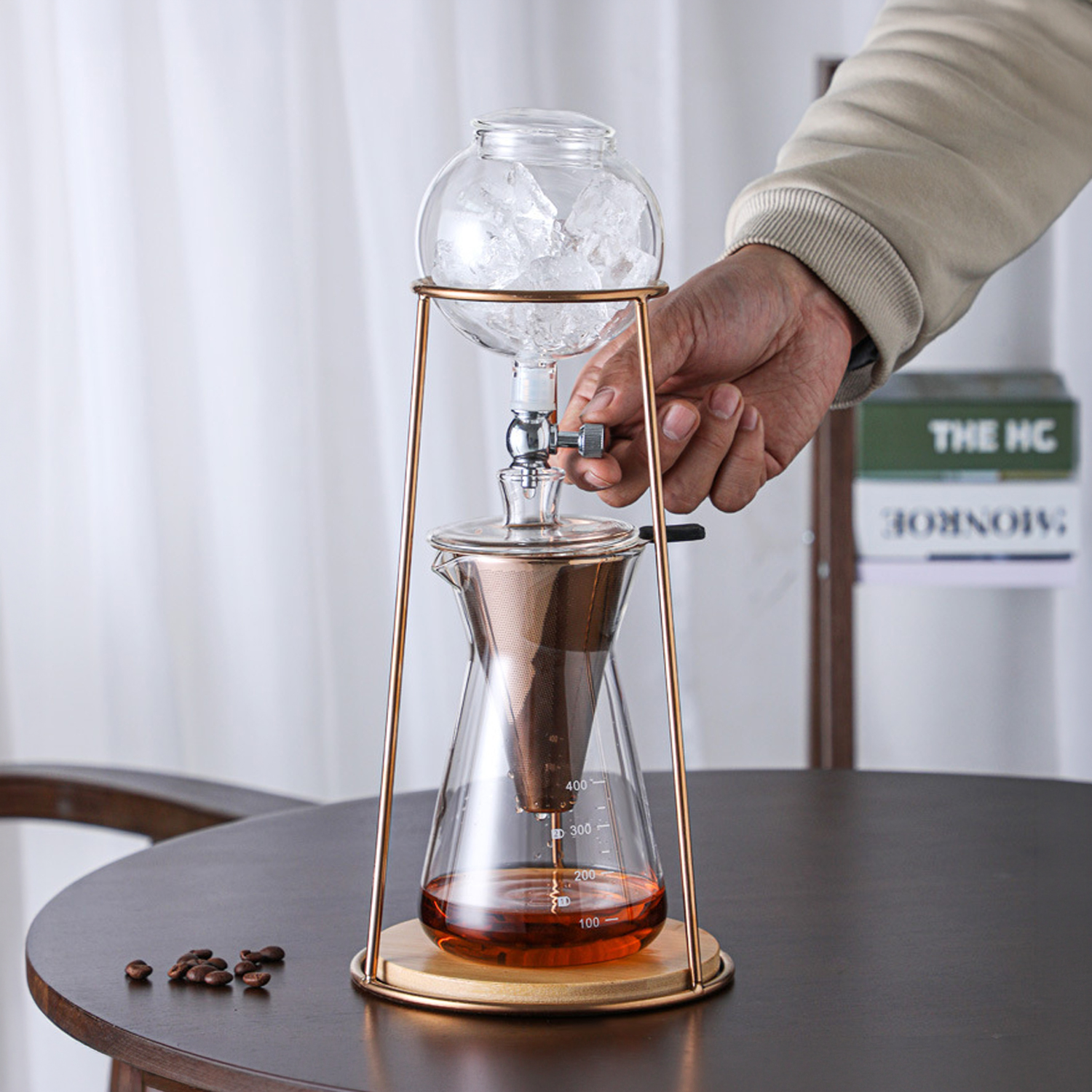 Una cafetera de goteo en frío de vidrio con hielo arriba y líquido abajo. Una mano ajusta una perilla.