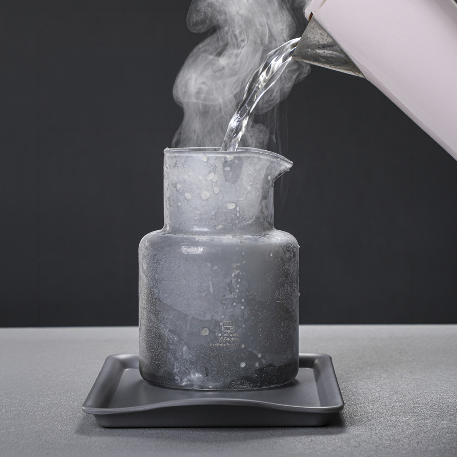 Se vierte agua caliente en un vaso escarchado, creando vapor. El vaso está sobre una bandeja oscura.