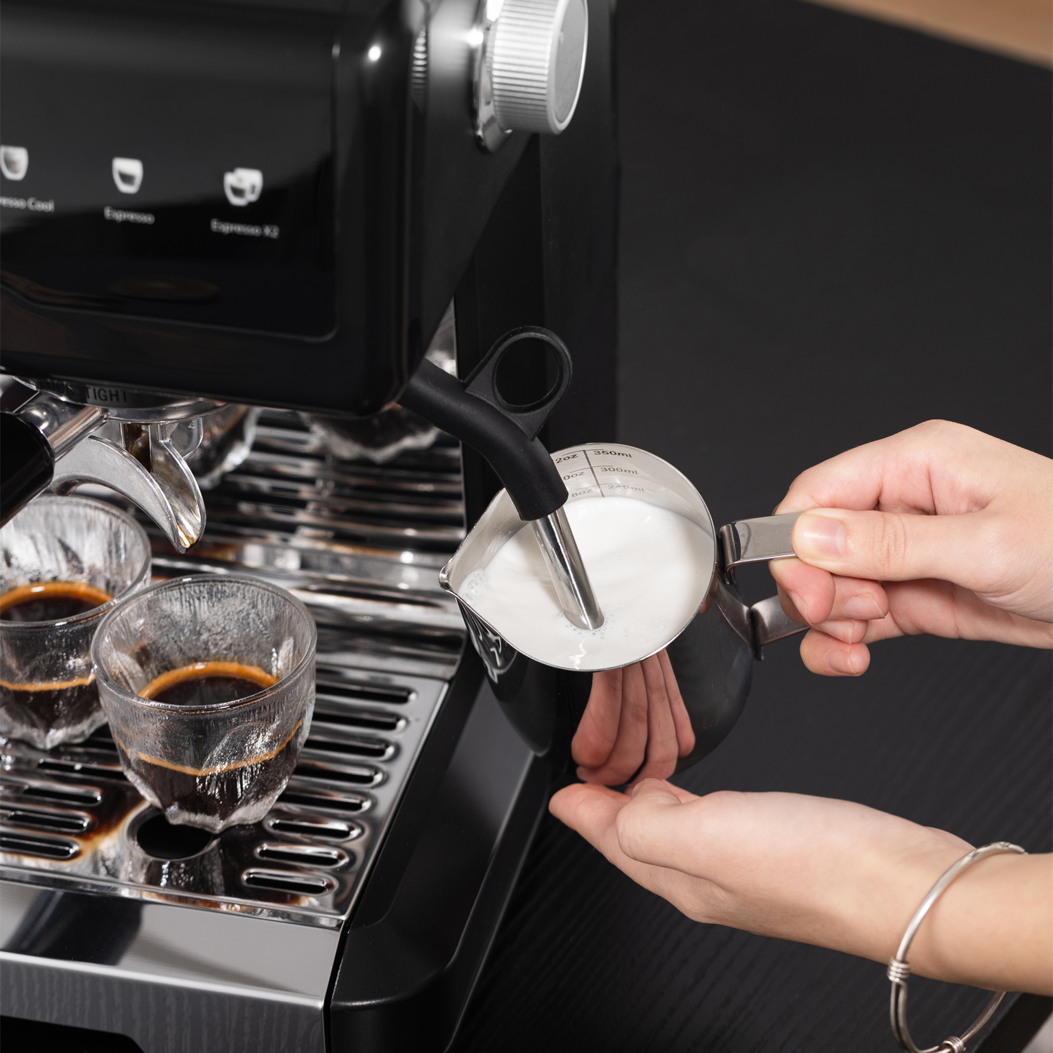 Eine Person schäumt Milch in einem Metallkännchen über einer Espressomaschine mit zwei Espressotassen.