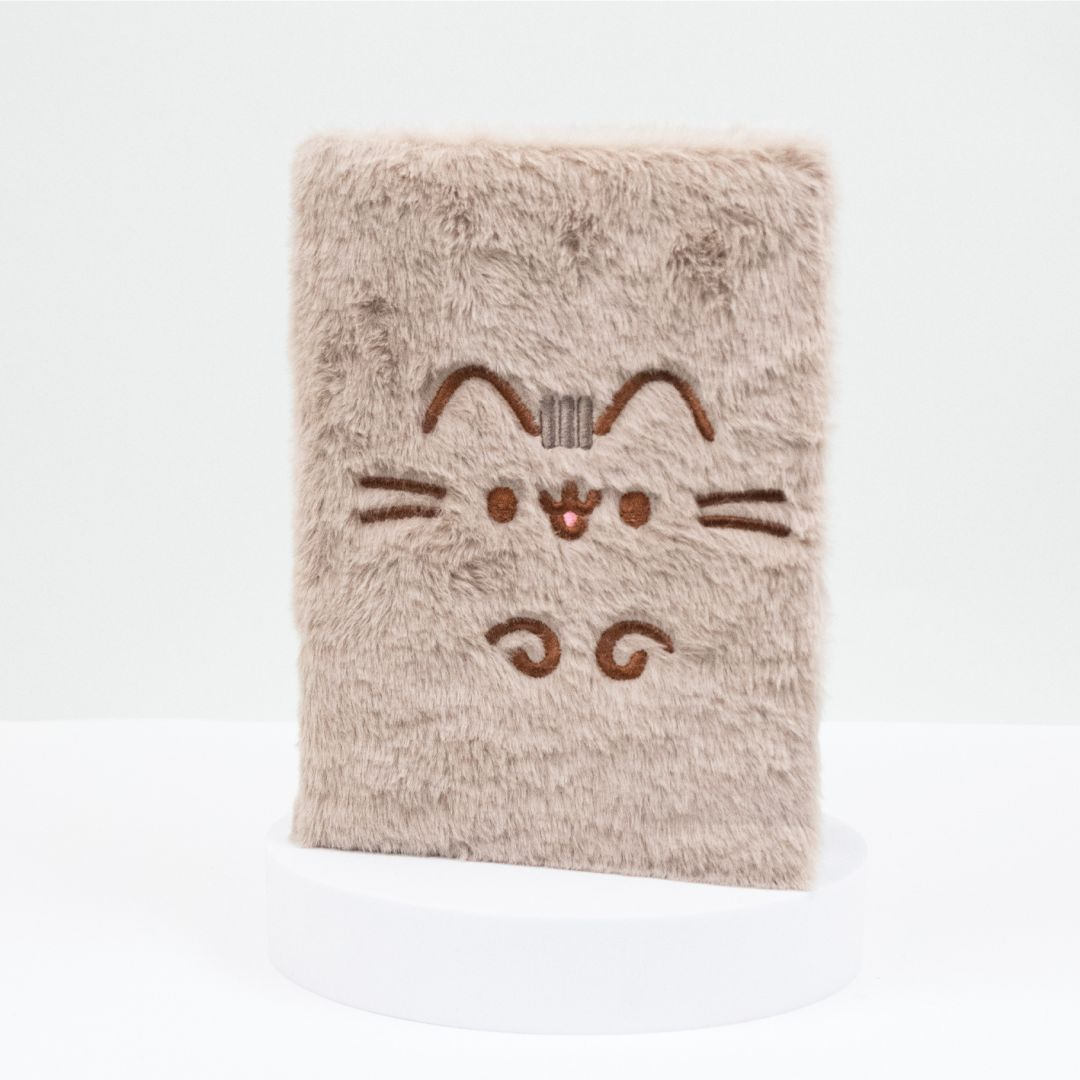 Un carnet beige pelucheux avec un visage de chat brodé.