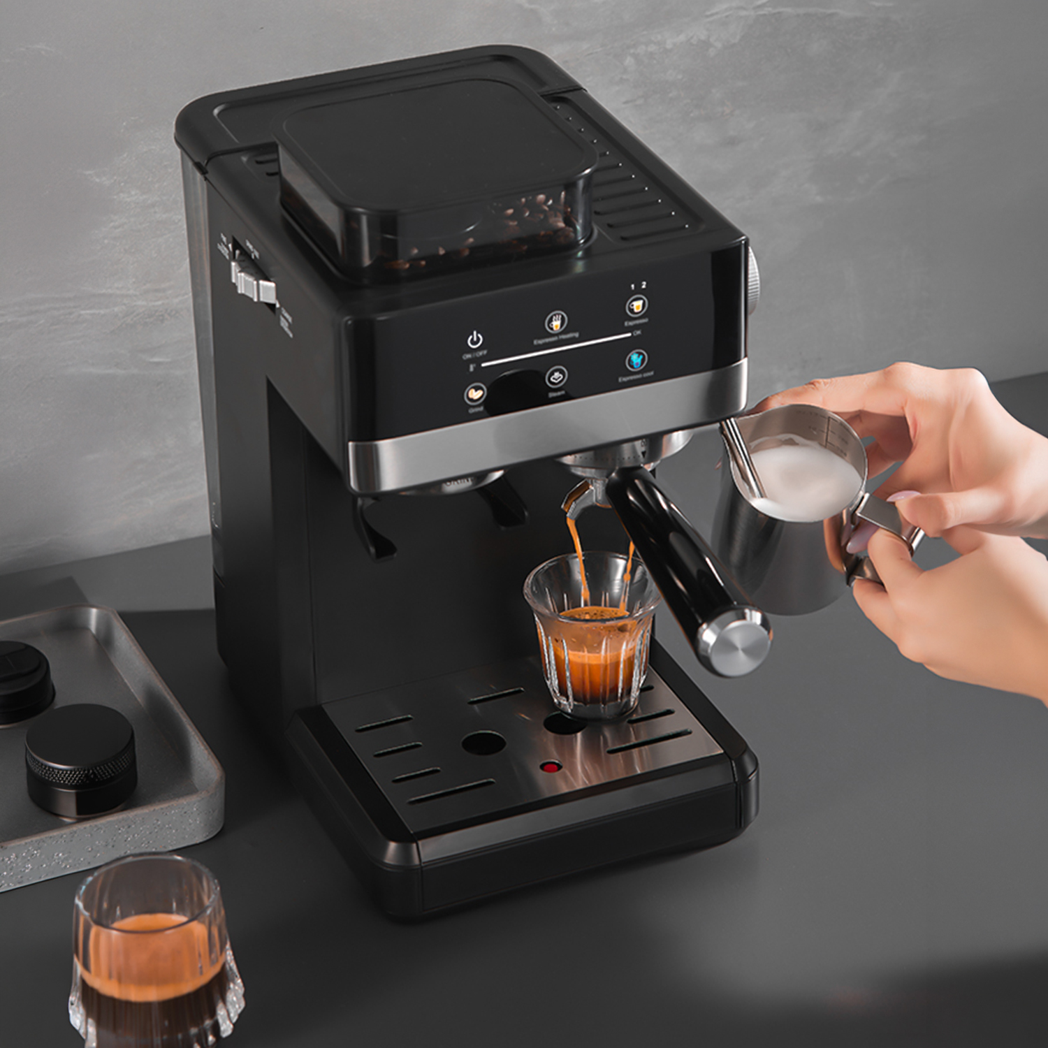 Eine schwarze Kaffeemaschine brüht Espresso in ein Glas, Milchschaum wird daneben vorbereitet.