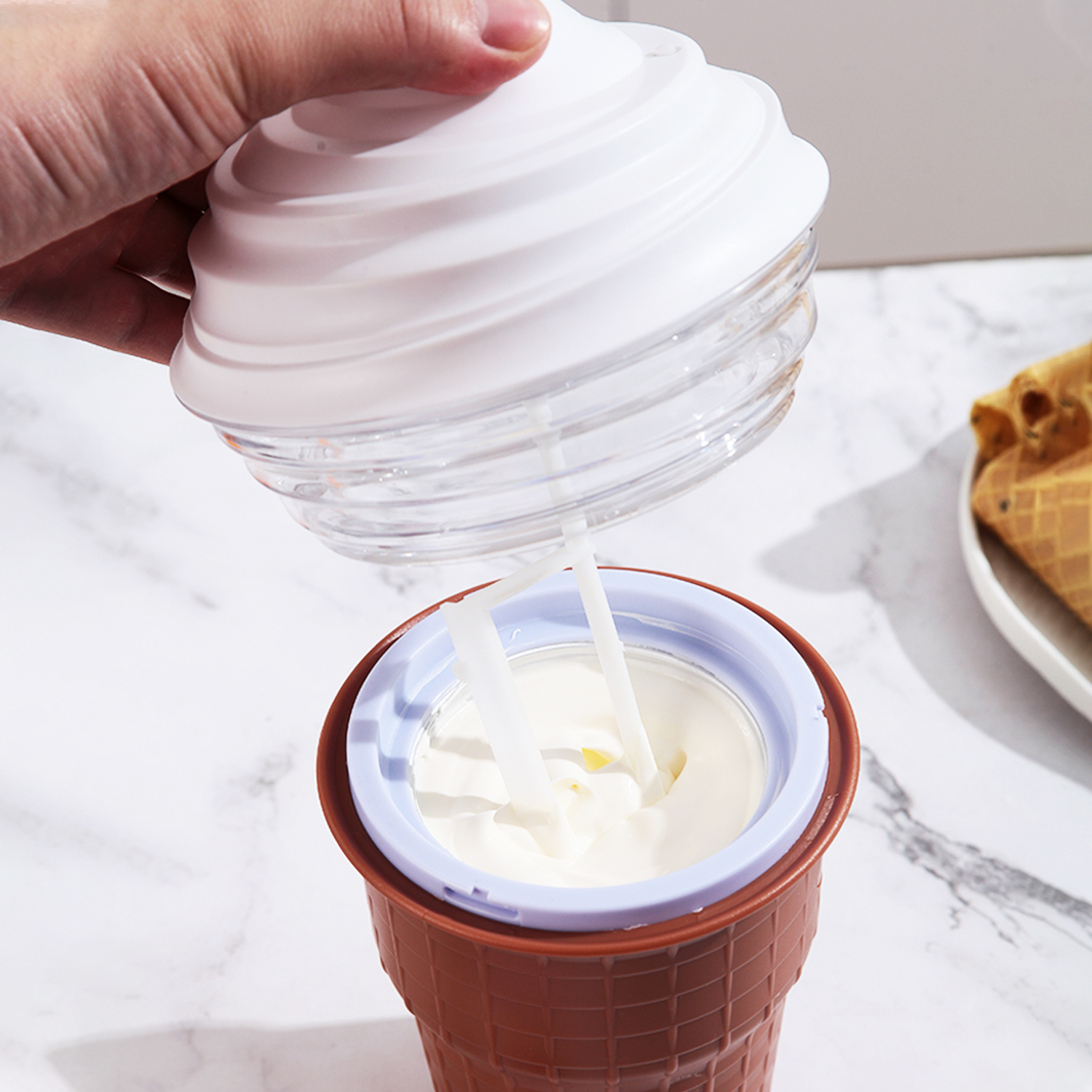 Hand hält Deckel einer Eismaschine mit weißer Creme in braunem Waffelbecher.
