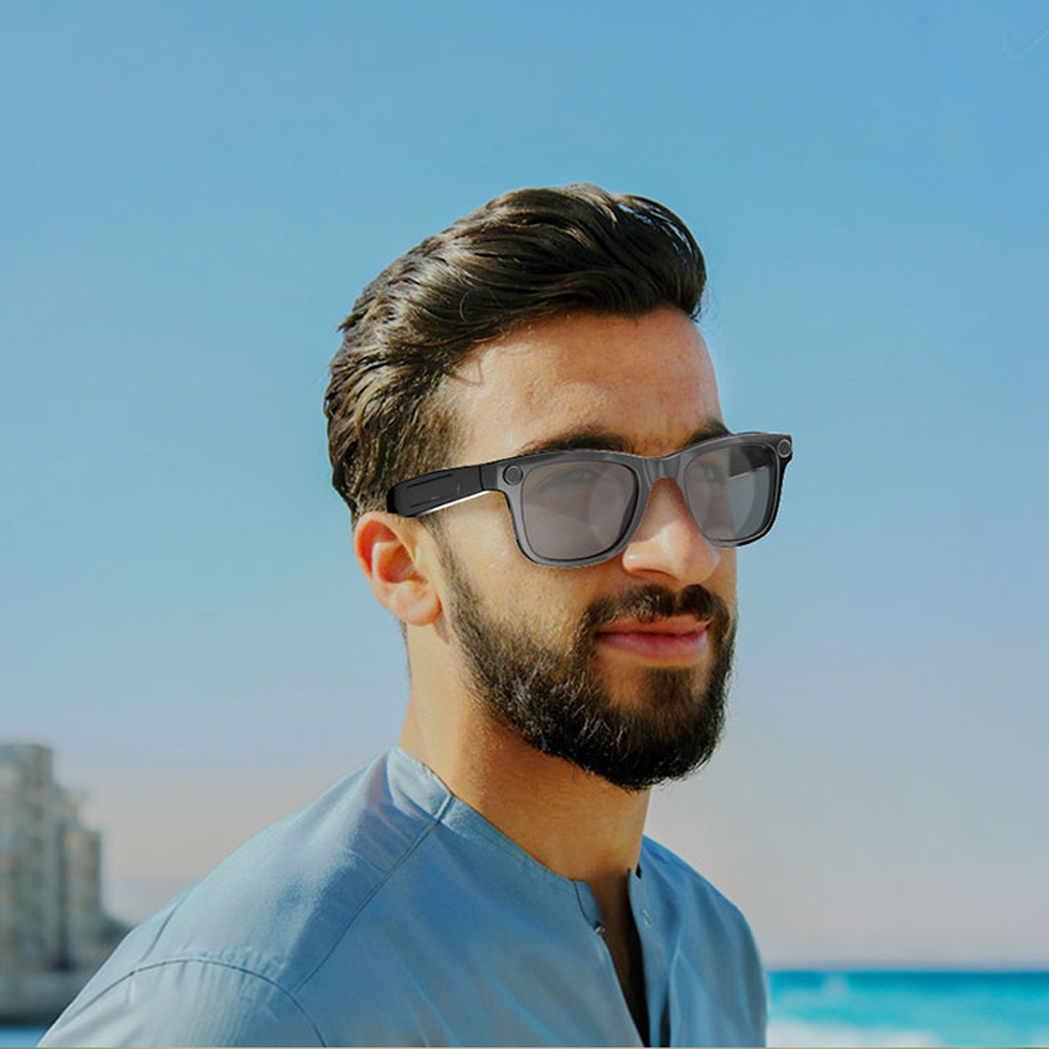 Mann mit Sonnenbrille am Strand, blauer Himmel, Meer und Gebäude im Hintergrund.