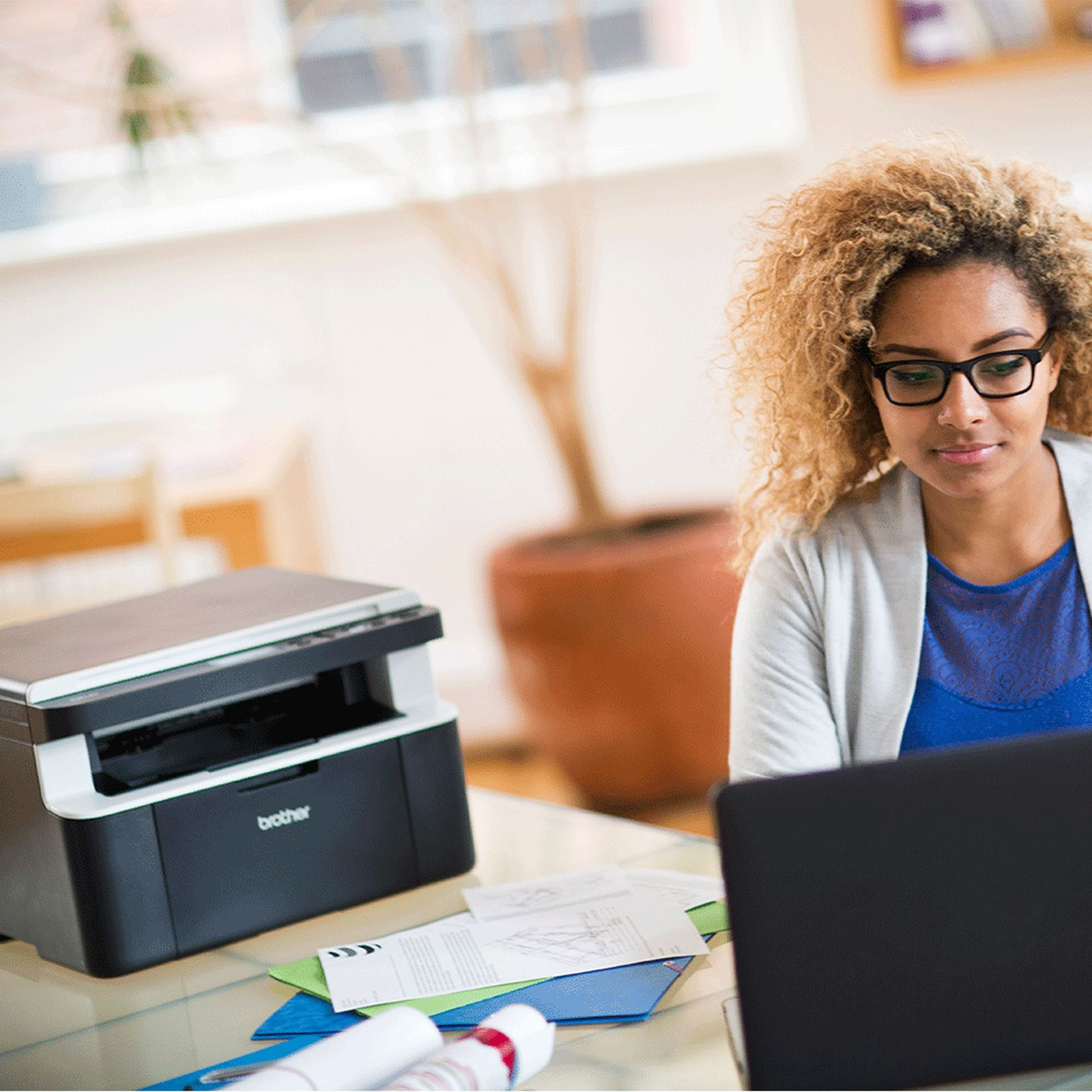 Eine Frau mit lockigem Haar und Brille arbeitet an einem Laptop neben einem Drucker.