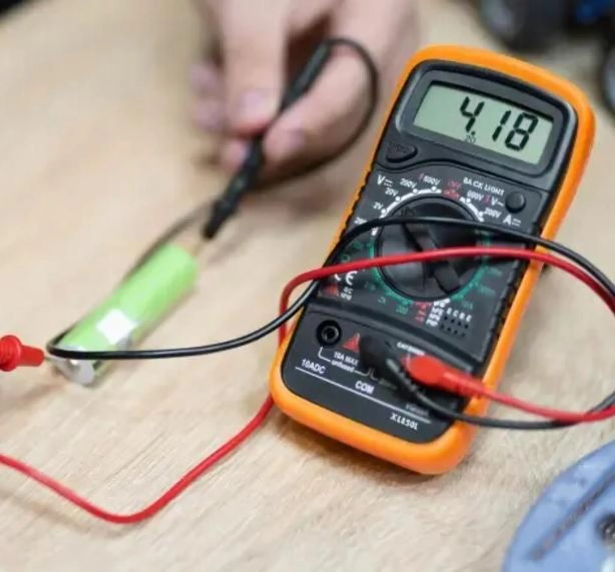 Ein orangefarbenes Multimeter misst eine grüne Batterie mit roten und schwarzen Kabeln.