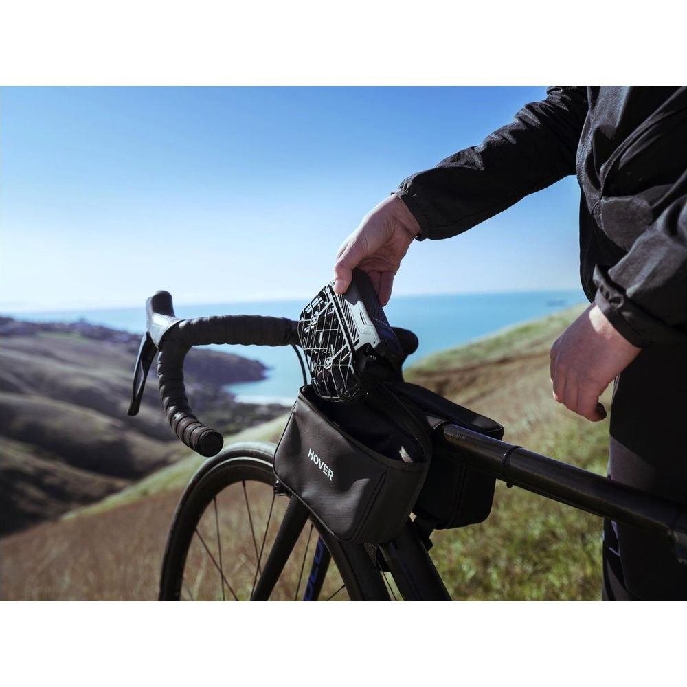 Manillar de bicicleta con bolsa, persona sujetando dron, paisaje montañoso, mar.