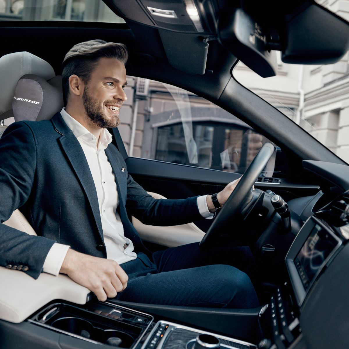A man in a suit drives a car, smiling.
