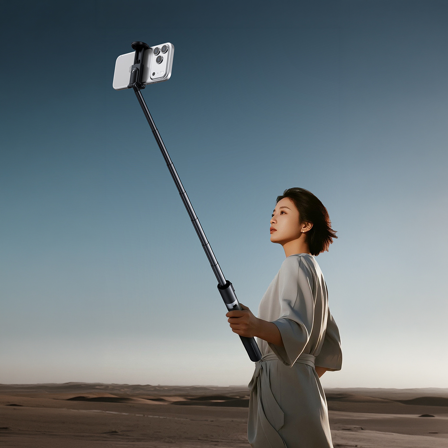 Eine Frau in einem grauen Kleid hält einen Selfie-Stick mit einem Handy in der Wüste.