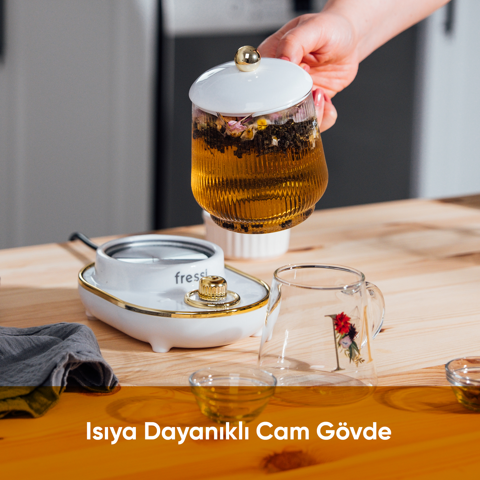 Bir el, çay ve çiçeklerle dolu, altın kapaklı cam demlik tutuyor. Yanında ısıtıcı ve bardak var.