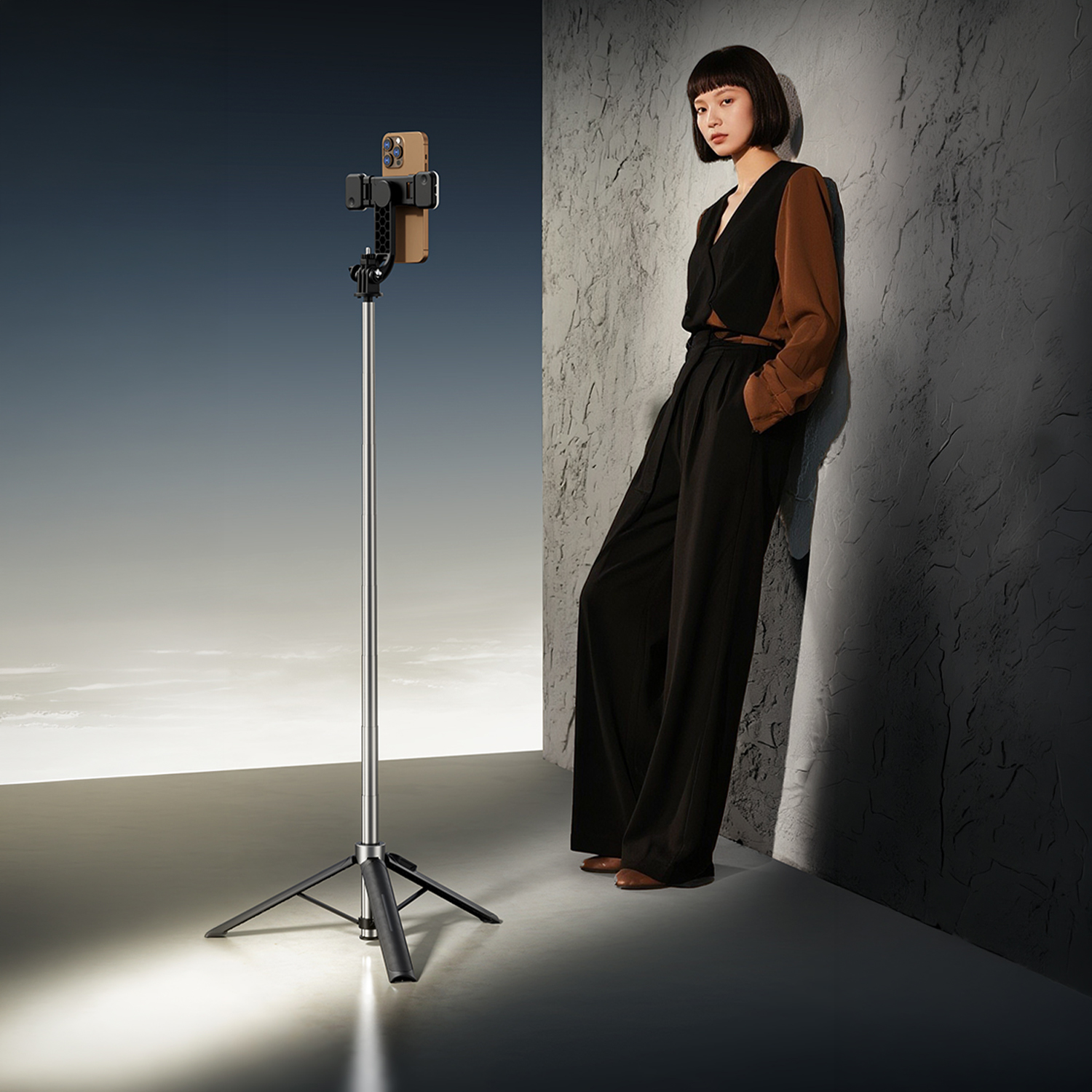 Eine Frau in einem schwarz-braunen Outfit lehnt an einer strukturierten Wand neben einem Stativ mit Handy.