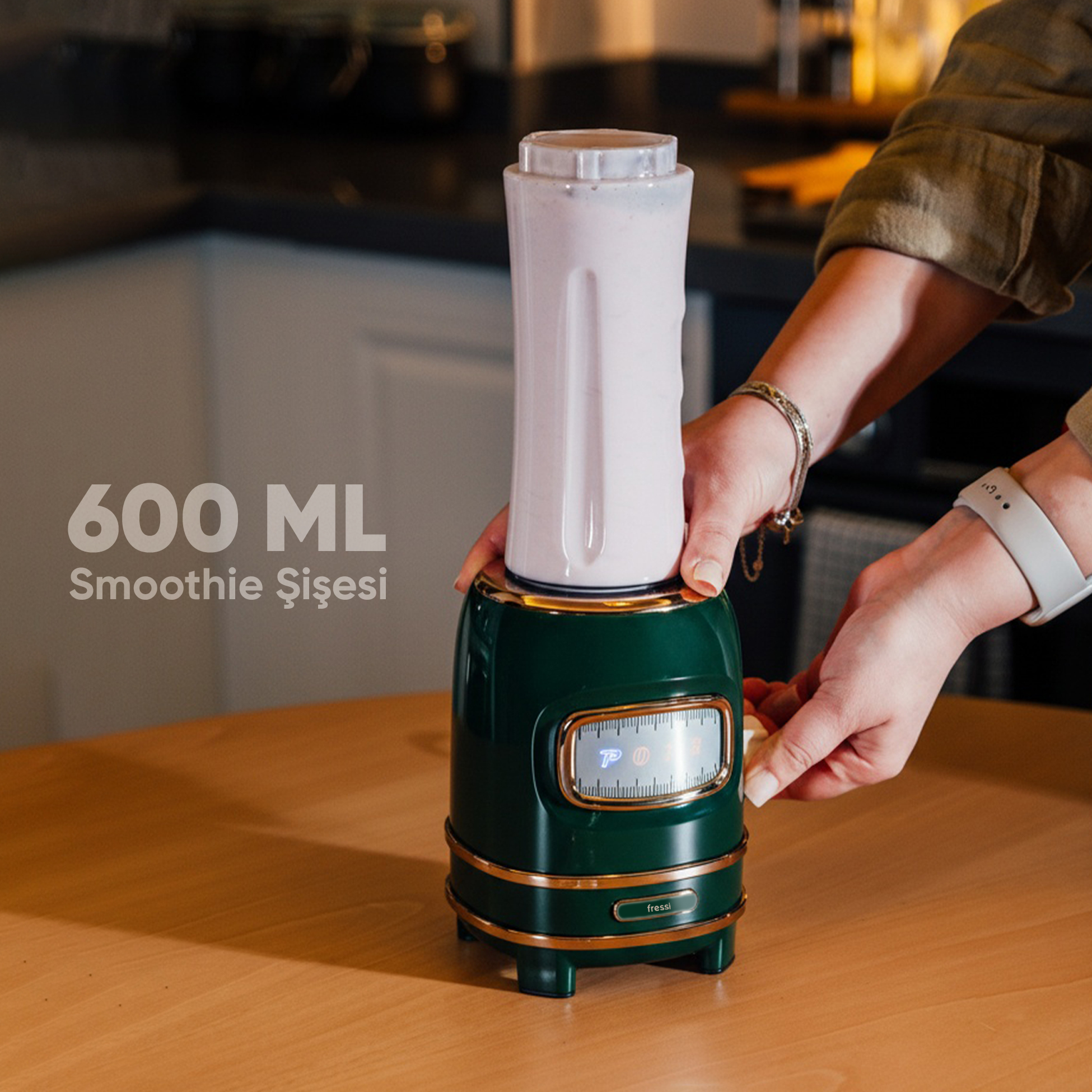 Koyu yeşil, beyaz haznelı ve altın detaylı bir blenderı bir kişi kullanıyor.