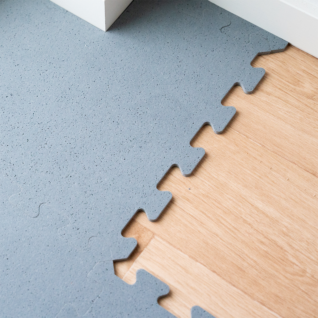 Graue Puzzlematten liegen auf einem Holzboden, daneben eine weiße Leiste.