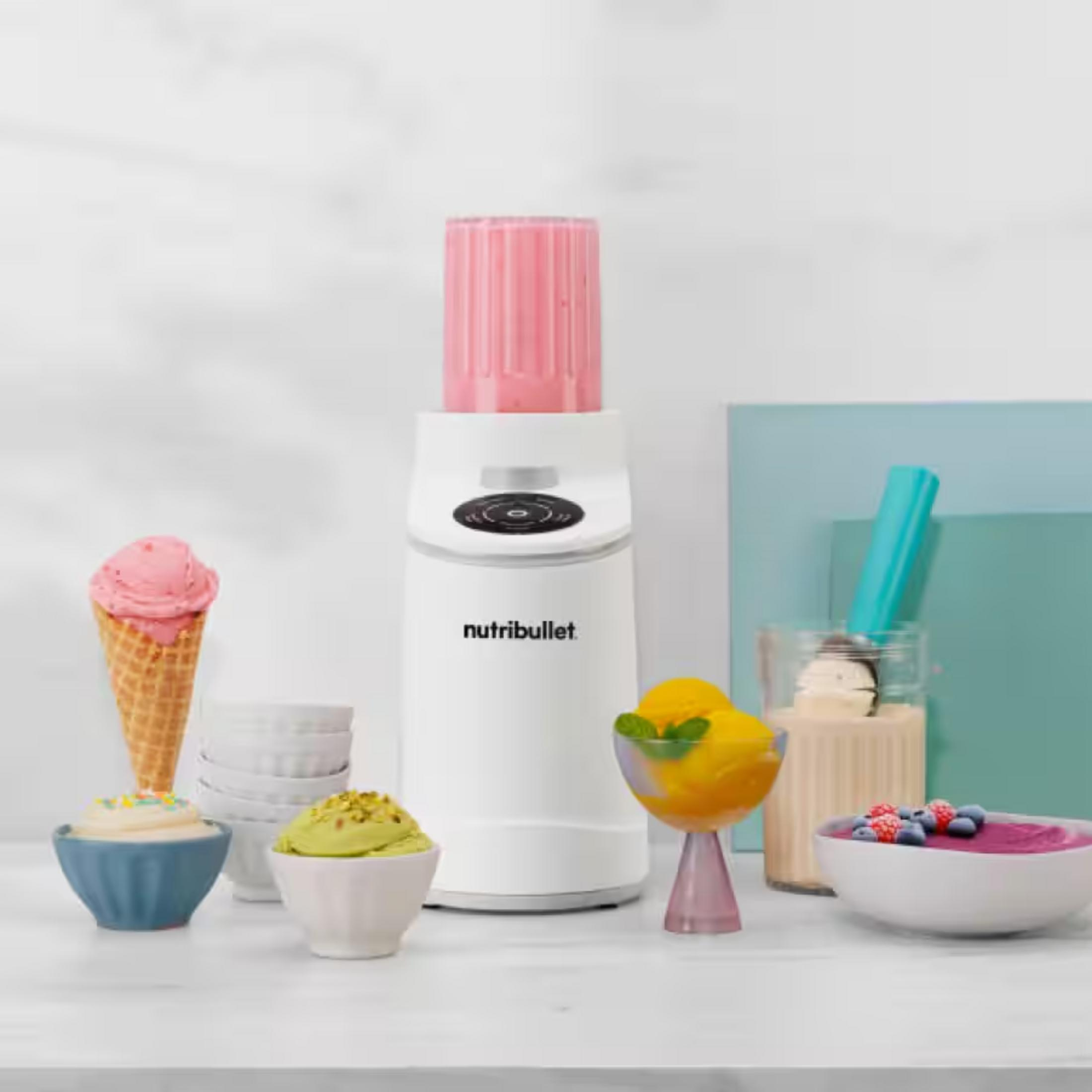 Ein weißer Nutribullet Mixer mit Eiscreme, Beeren und Obstschalen auf einem Tisch.
