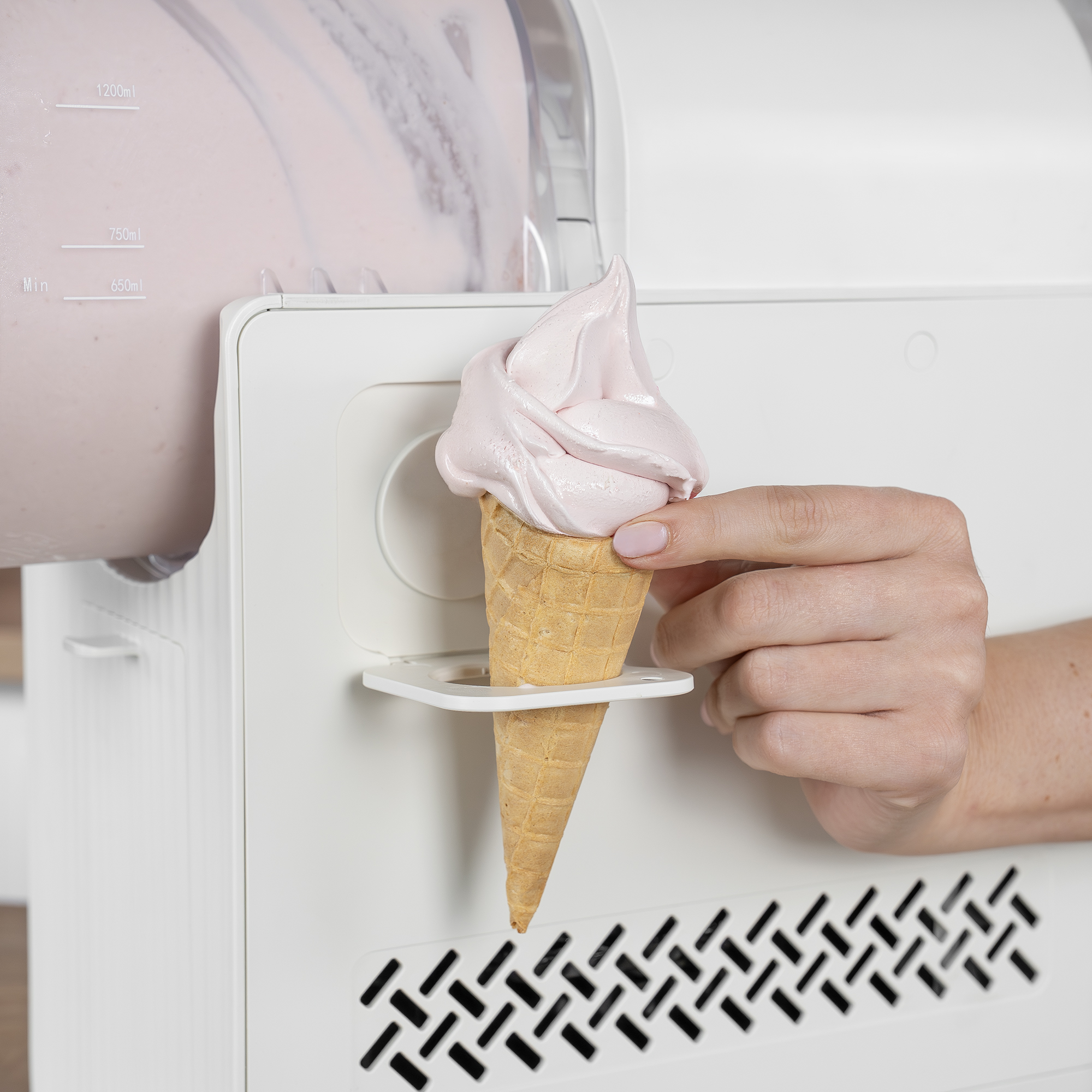 Una mano tiene un cono gelato con gelato soft rosa sotto un erogatore di gelatiera.