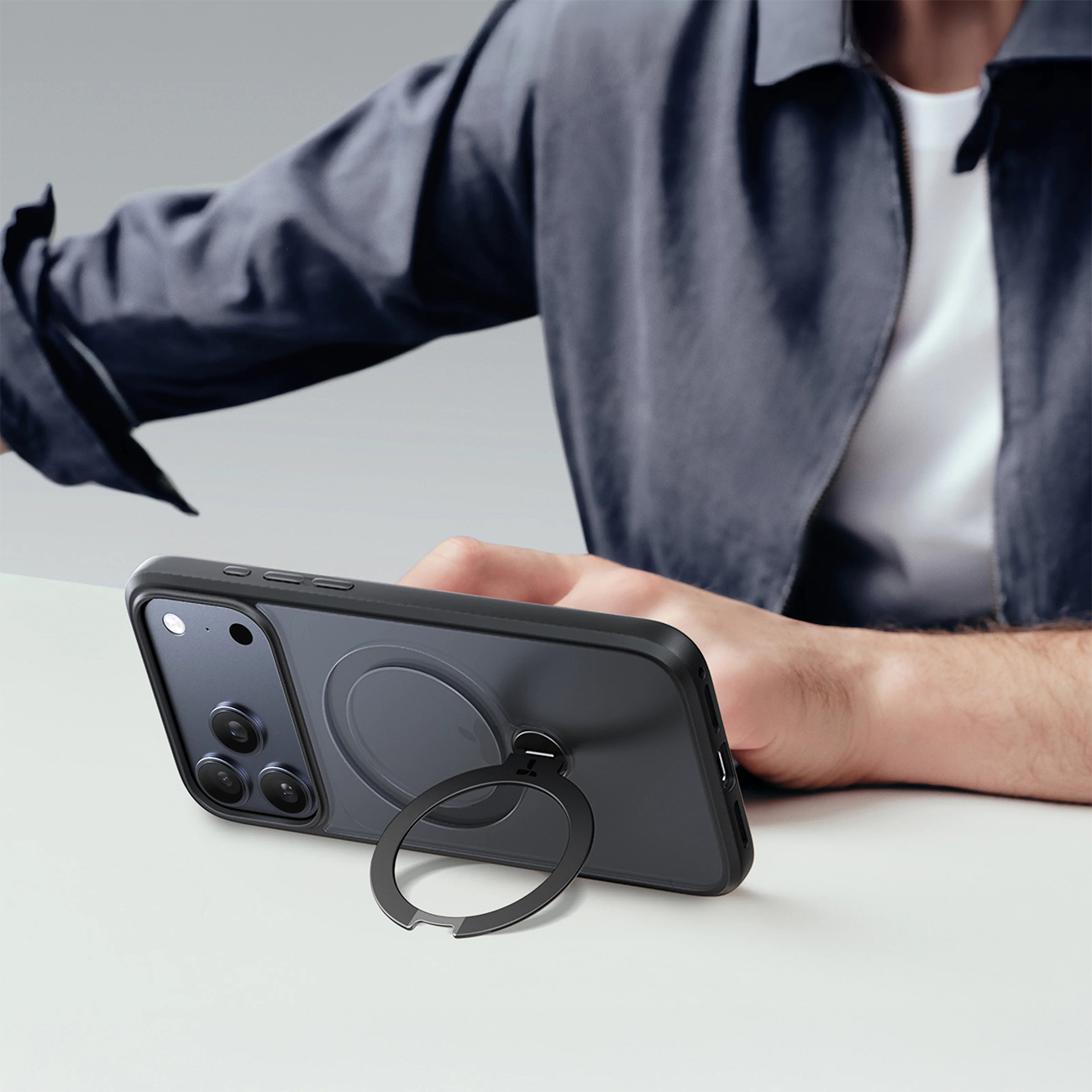 Eine Hand hält ein schwarzes iPhone mit Ringhalterung. Eine Person im dunklen Jackett ist unscharf.