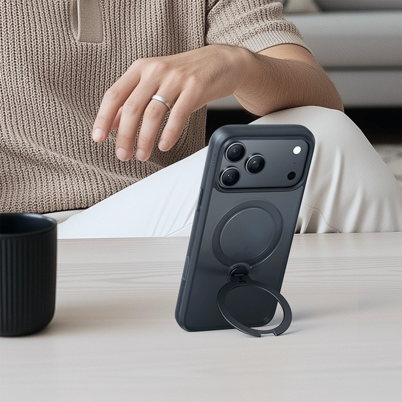 Ein dunkles Handy mit Ringständer steht neben einer schwarzen Tasse auf einem Holztisch.
