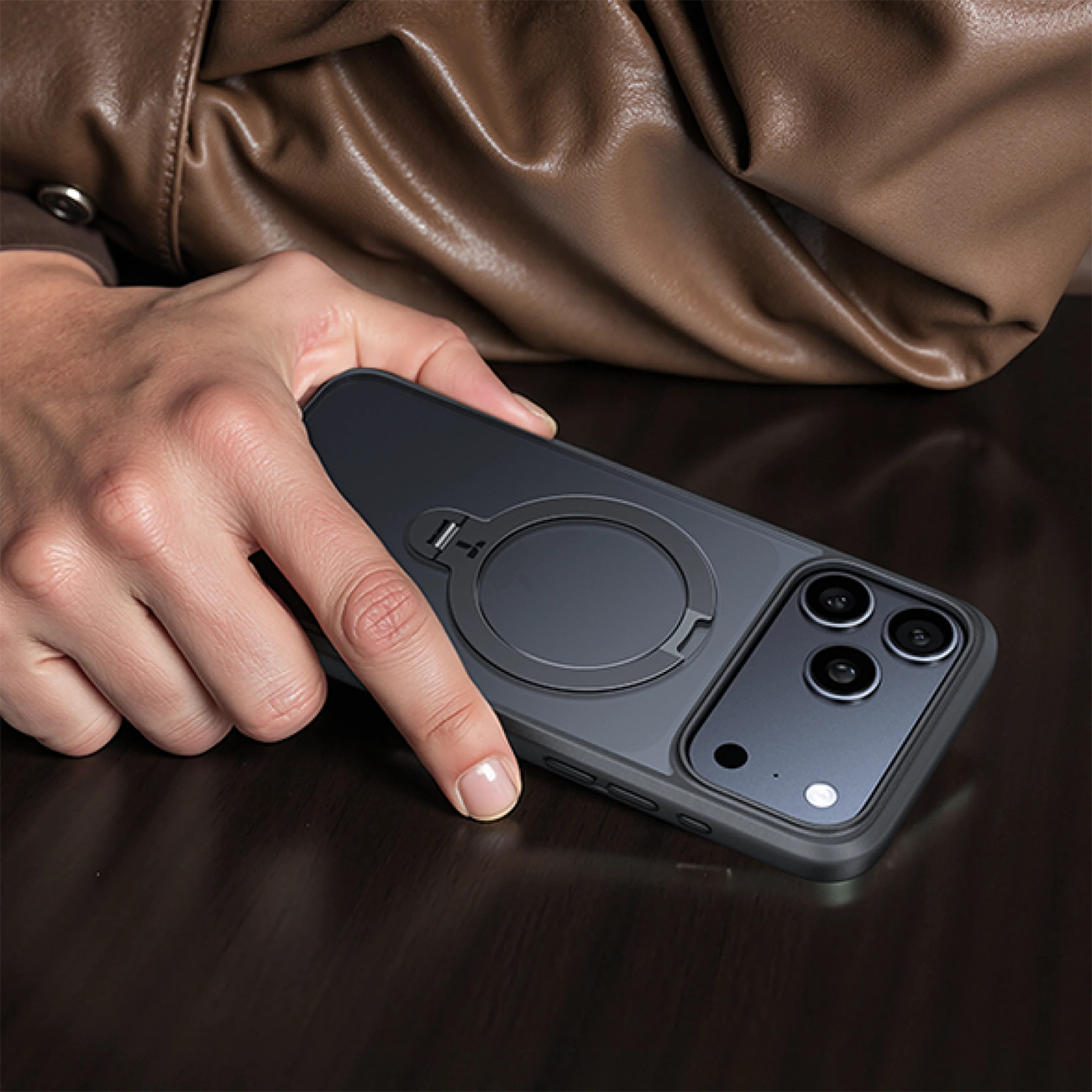 Eine Hand hält ein schwarzes Handy mit Ringhalterung vor brauner Lederjacke.