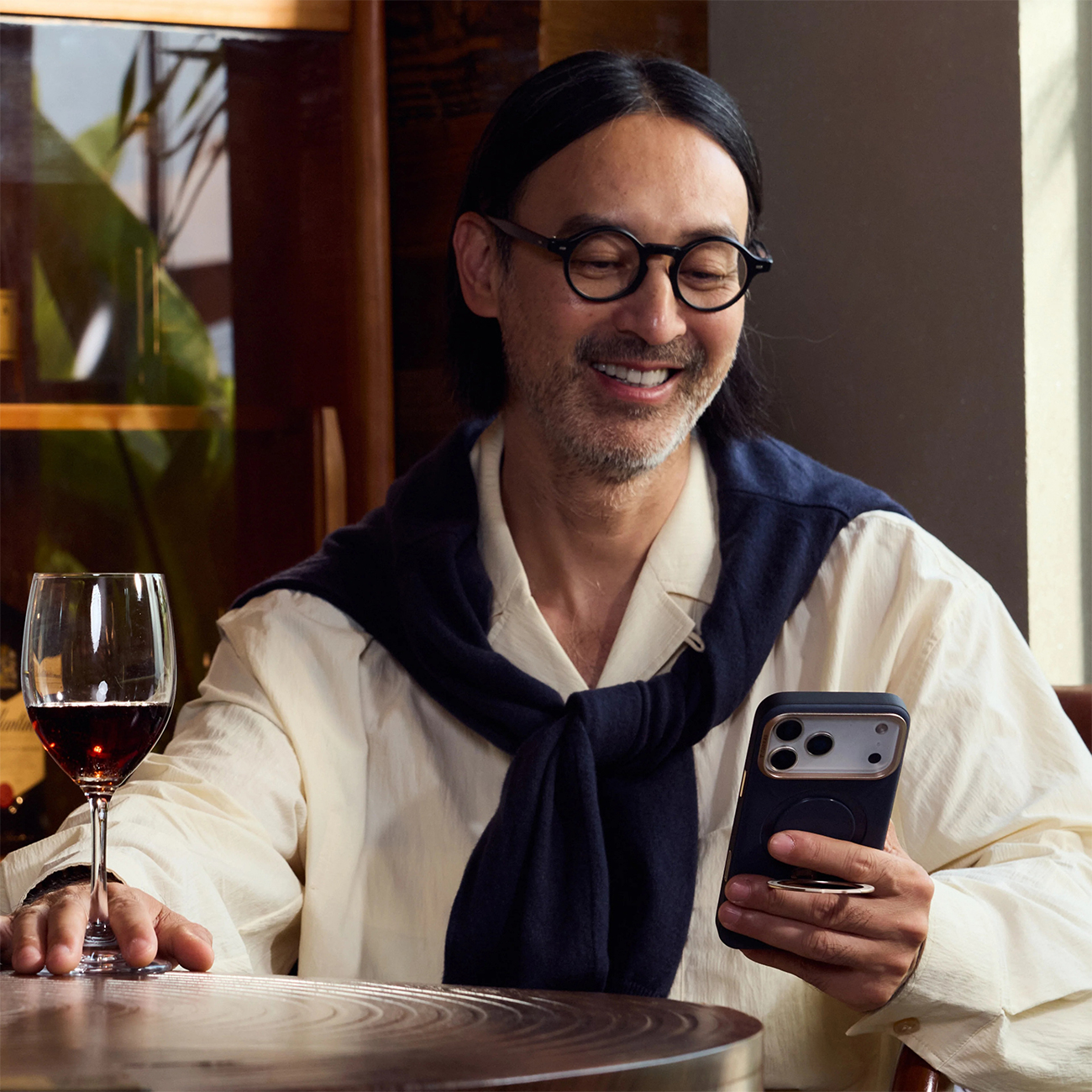Un hombre con gafas y suéter en los hombros sostiene un teléfono, junto a una copa de vino.
