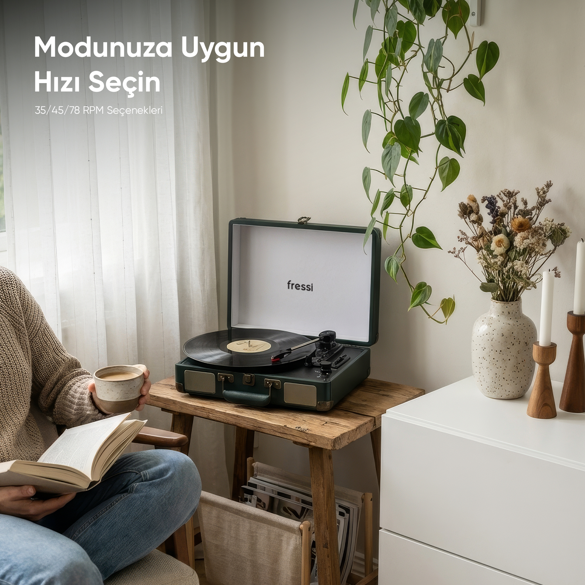 Bir kişi kitap okuyor ve bir pikap ile kuru çiçeklerin yanında kahve fincanı tutuyor.