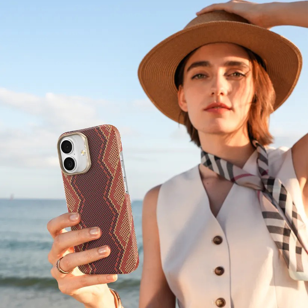 Mujer en la playa sujetando sombrero y teléfono. Mar y cielo de fondo.