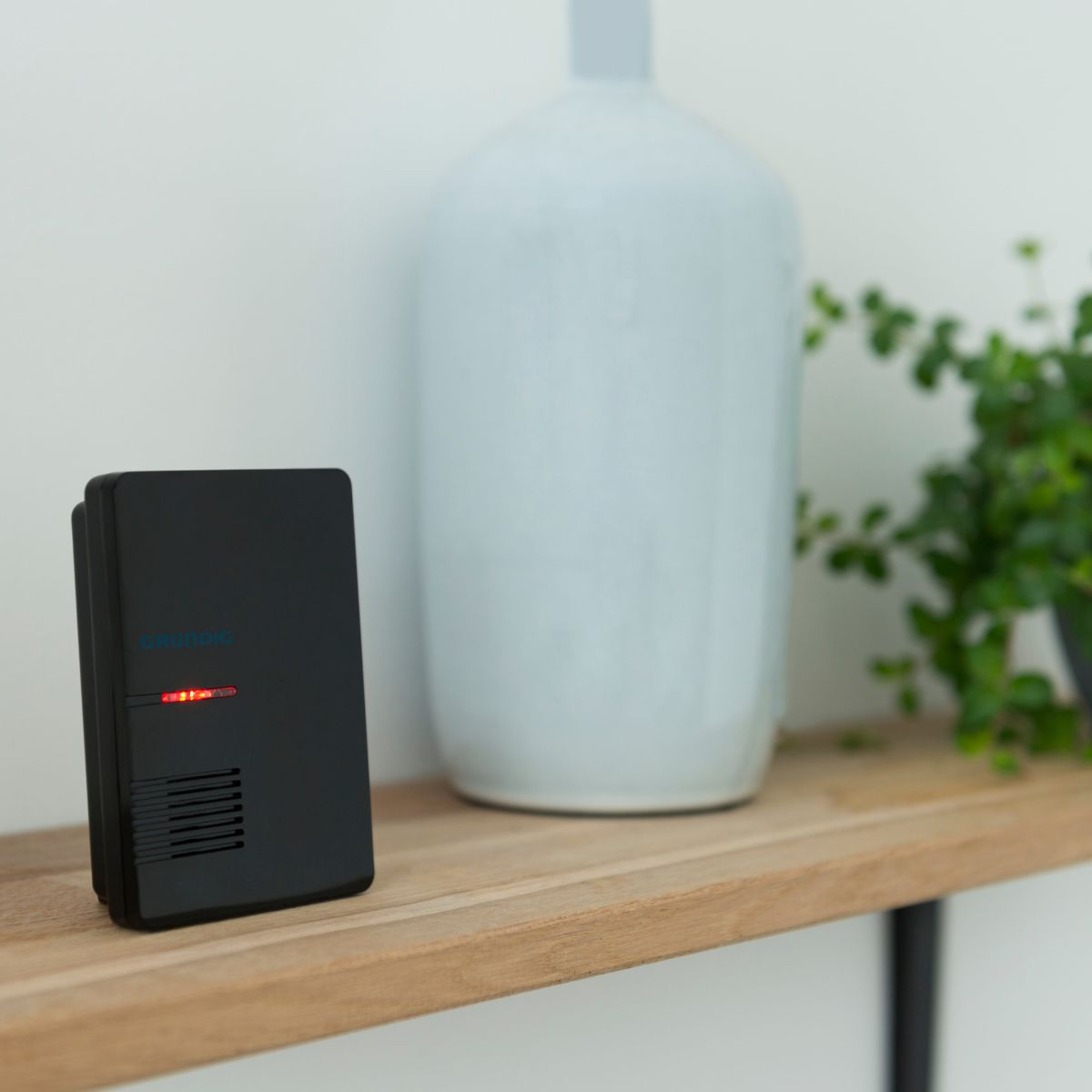 Un appareil noir avec une LED rouge sur une étagère en bois à côté d'un vase.