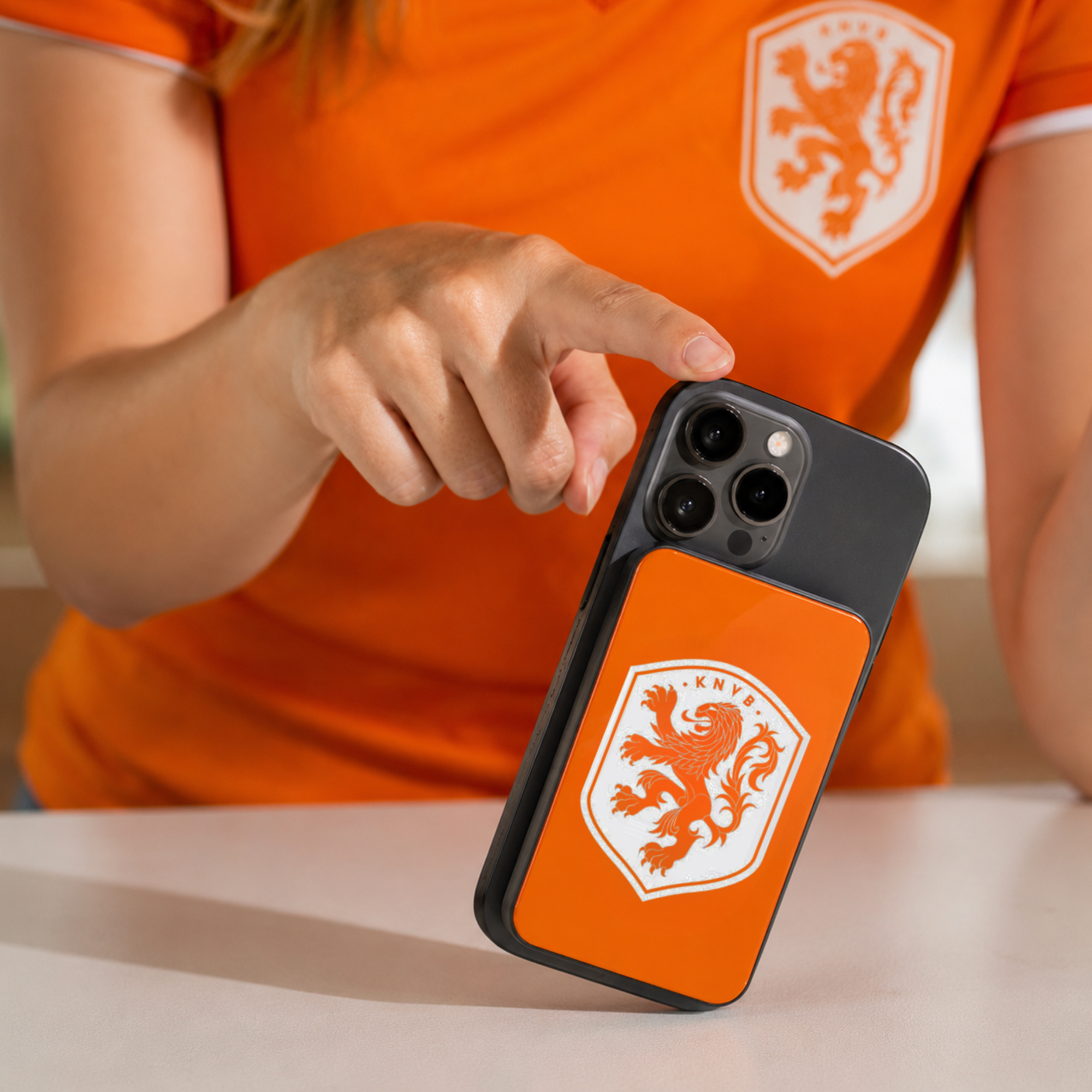 Persoon in oranje shirt met leeuwenembleem houdt telefoon met bijpassend embleem vast.