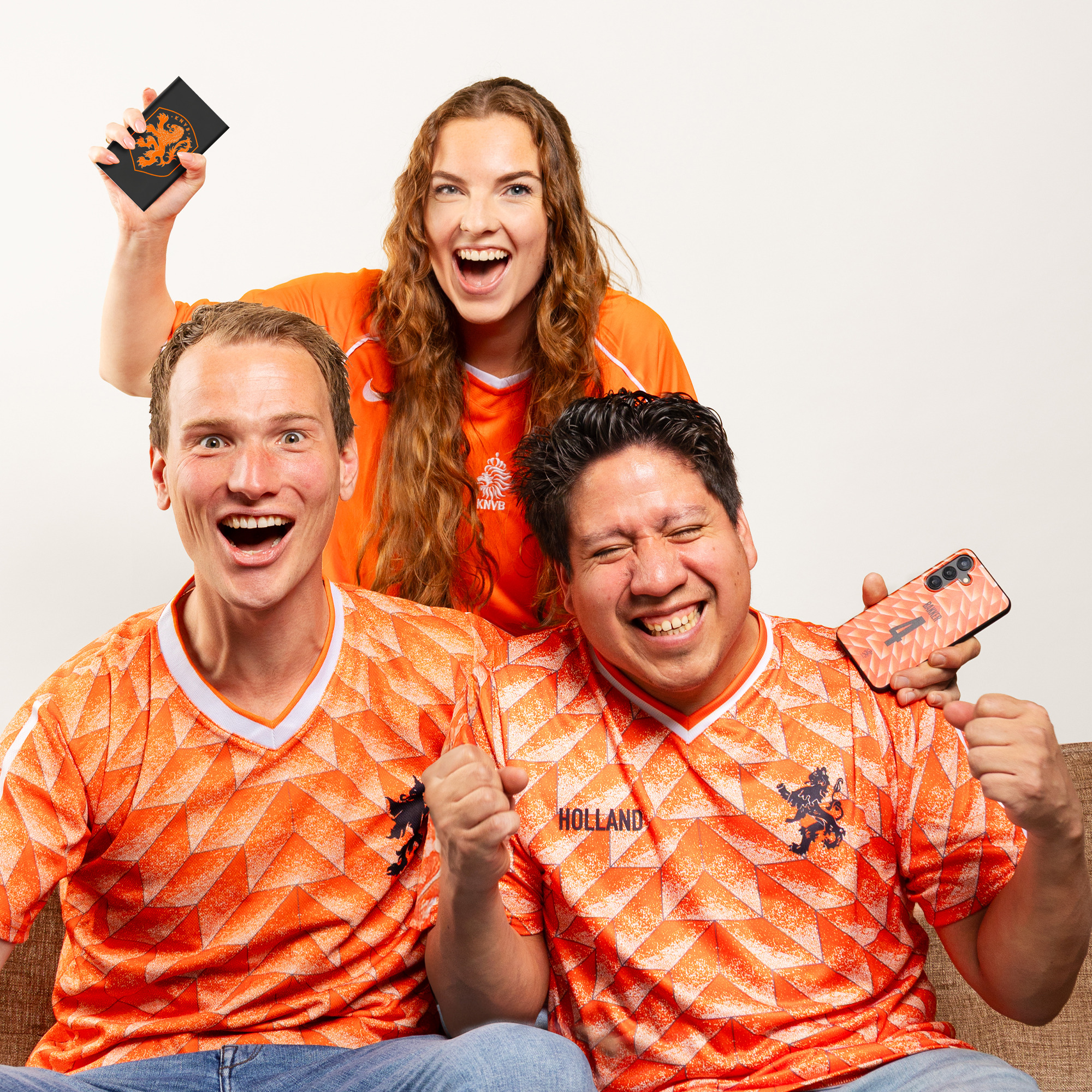 Drie fans in oranje shirts juichen. Eén houdt een telefoon vast.