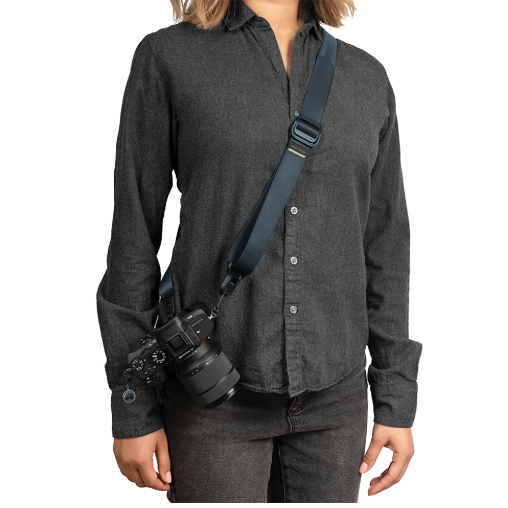 Persoon in donker shirt en jeans draagt een camera met blauwe riem.
