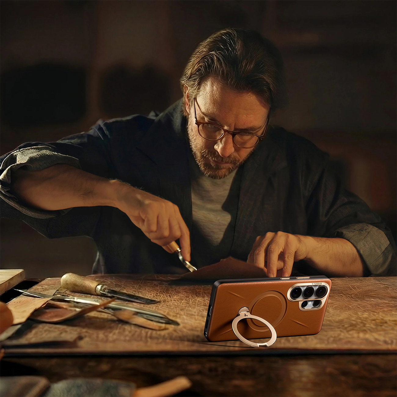 Hombre con gafas talla cuero con herramienta. Un teléfono yace a su lado.