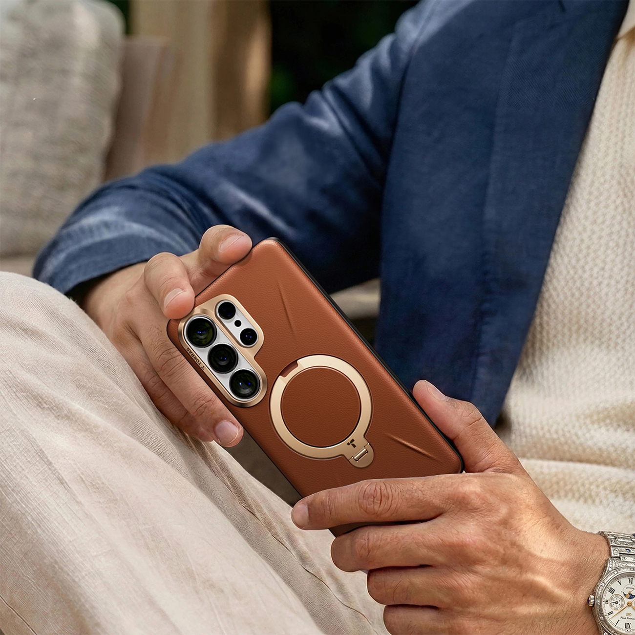 Hombre sosteniendo funda de teléfono marrón con soporte de anillo. Chaqueta azul, suéter beige.