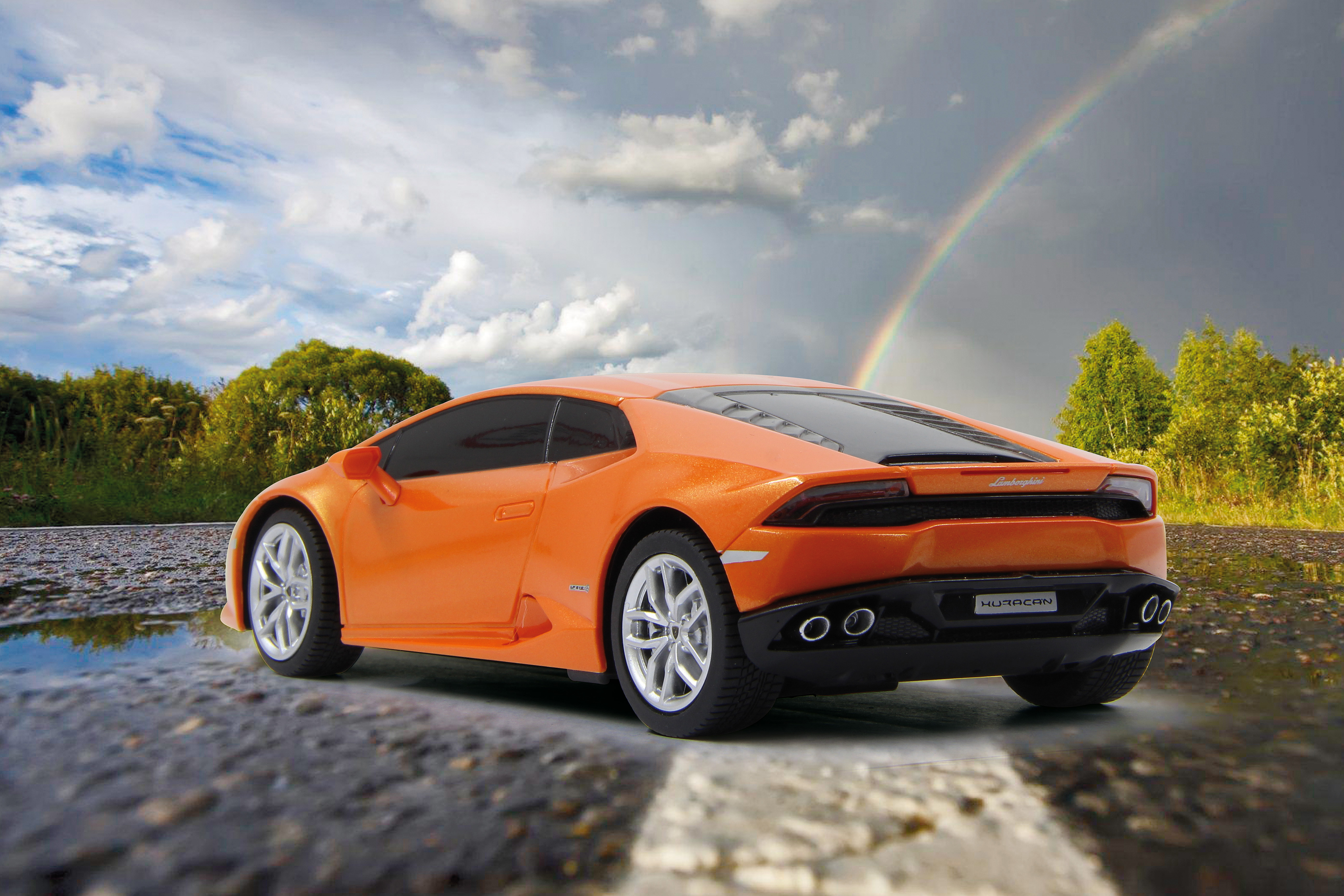 Ein oranges Spielzeugauto steht auf einer nassen Straße mit einem Regenbogen am Himmel.