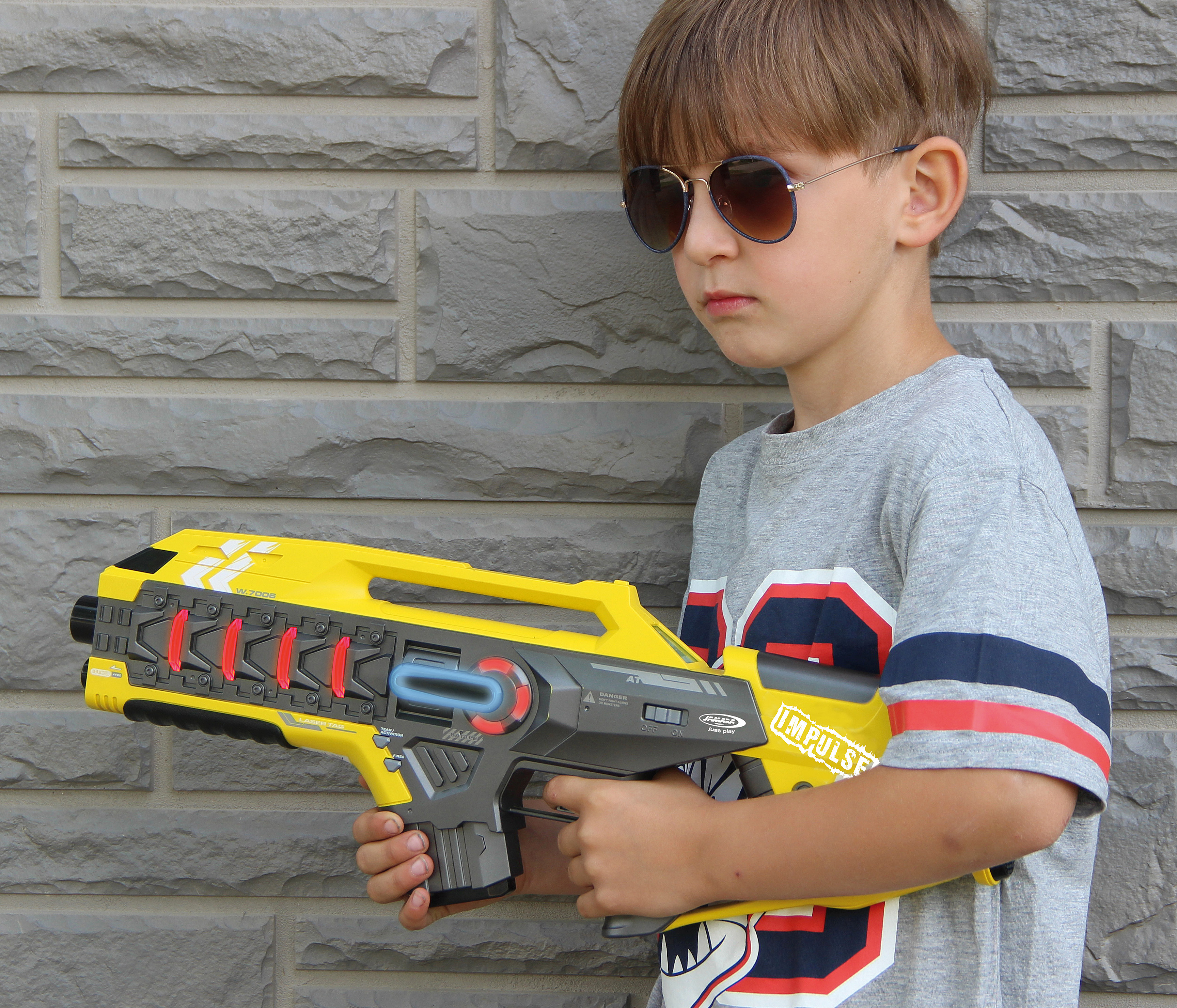 Ein Junge mit Sonnenbrille hält eine gelbe Laserpistole vor einer Backsteinwand.
